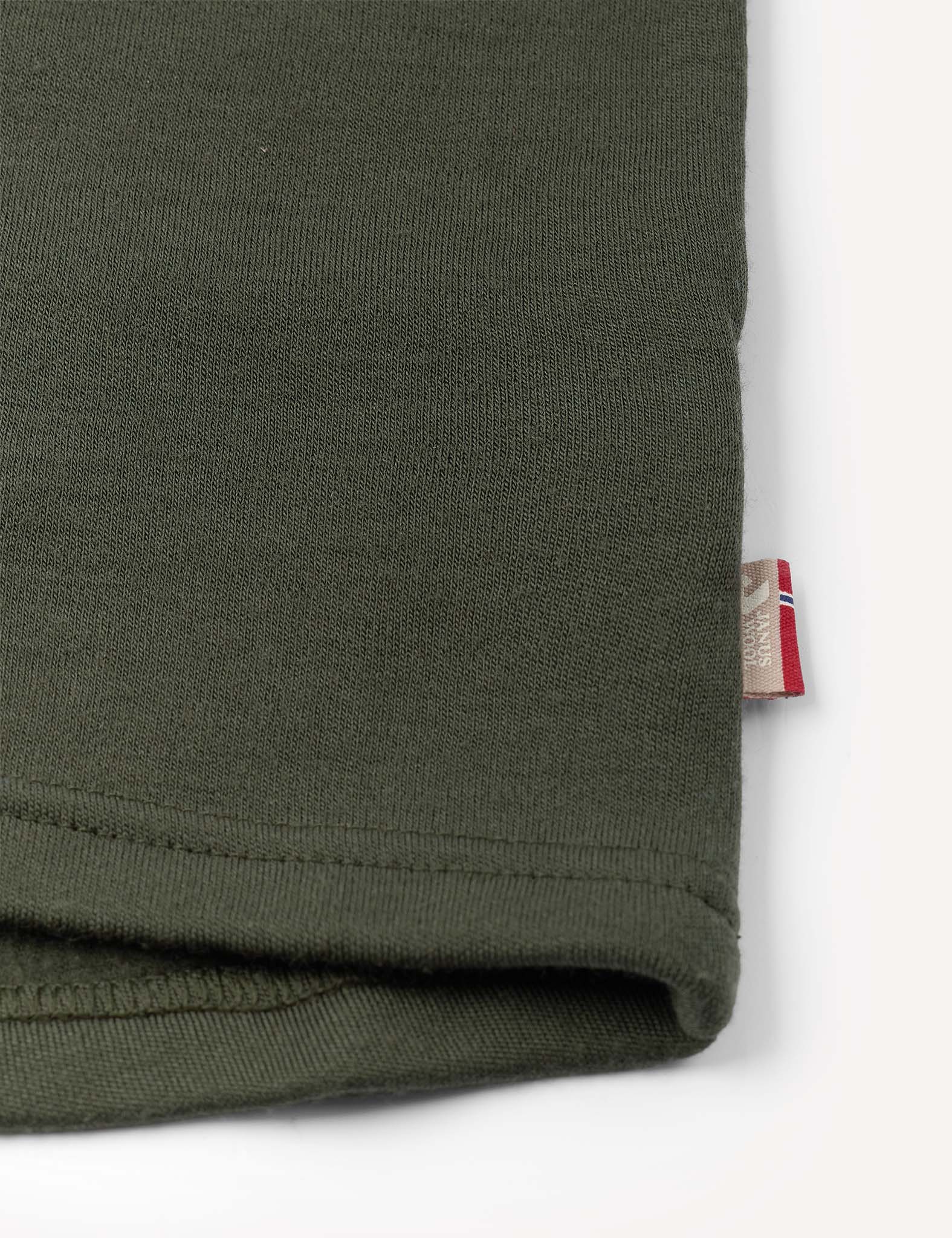 Close-up of the hem of the Crewneck – Merino Kids AirWool in olive green, featuring a small beige and red tag sewn into the seam, shown against a white background.