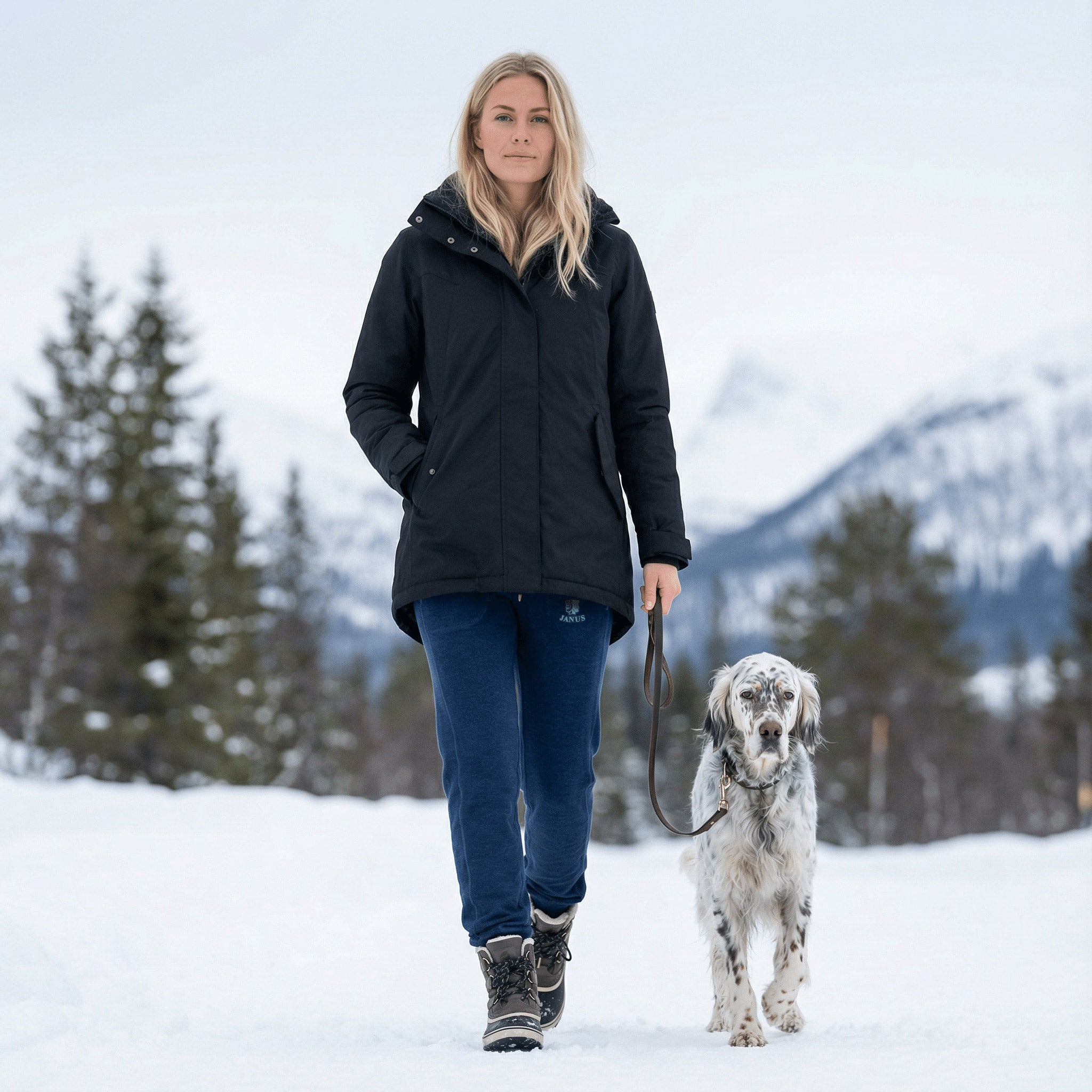 A woman in a black jacket and blue Pants merino - Designwool 350 Women by Janusfabrikken walks a white and brown dog on a leash through snow, surrounded by trees and mountains.