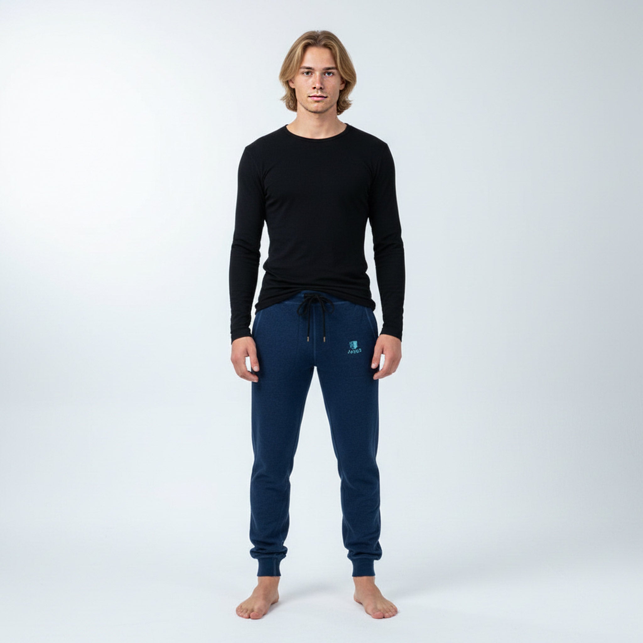 A person stands barefoot against a plain white background, wearing a fitted black long-sleeve shirt and dark blue Janus Wool Pants merino - Designwool 350 Men, facing the camera with arms relaxed at their sides.