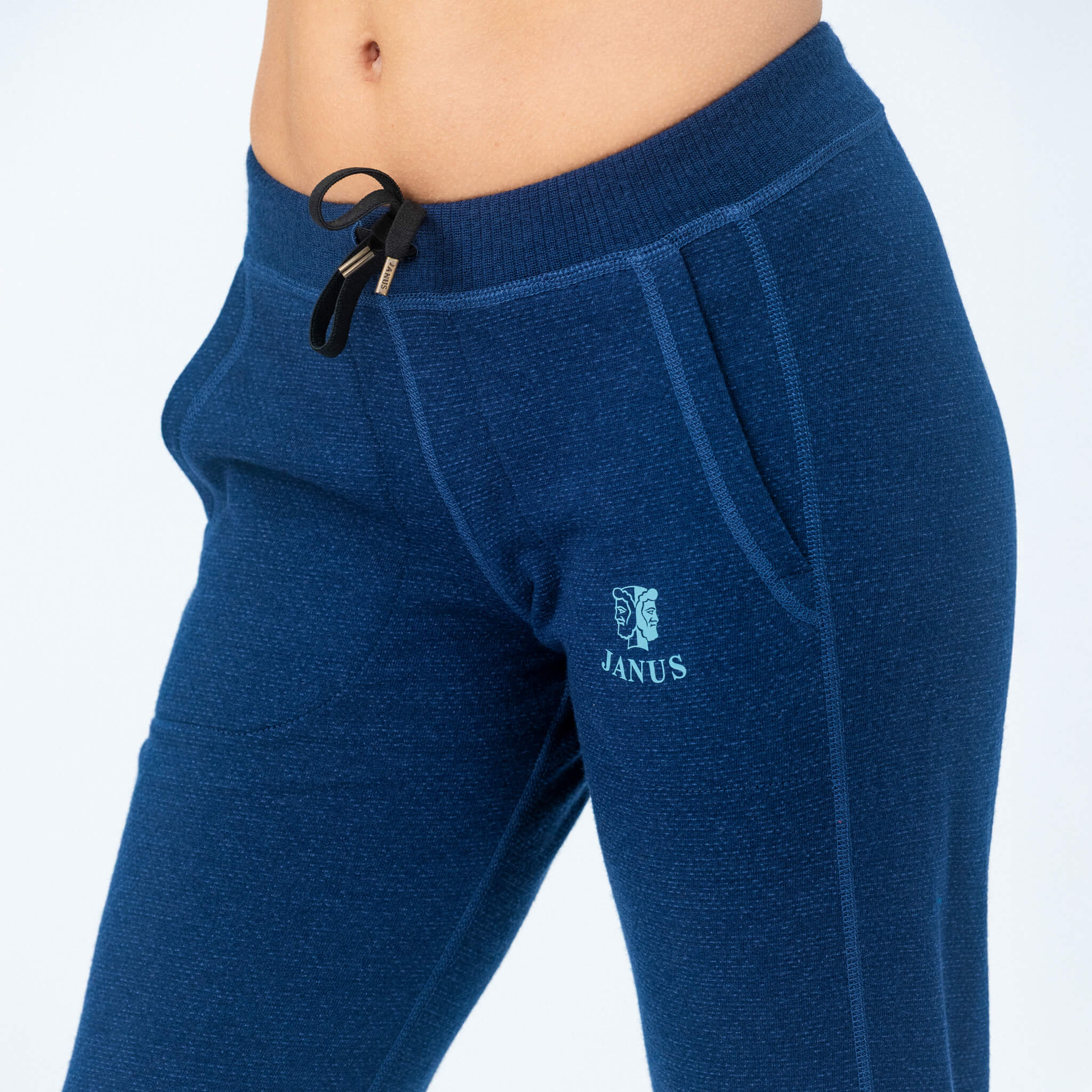 Close-up of a person wearing Janus Wool Pants merino - Designwool 350 Women, featuring pockets, a drawstring waist, and a logo on the left hip. The midriff is visible against a plain white background.