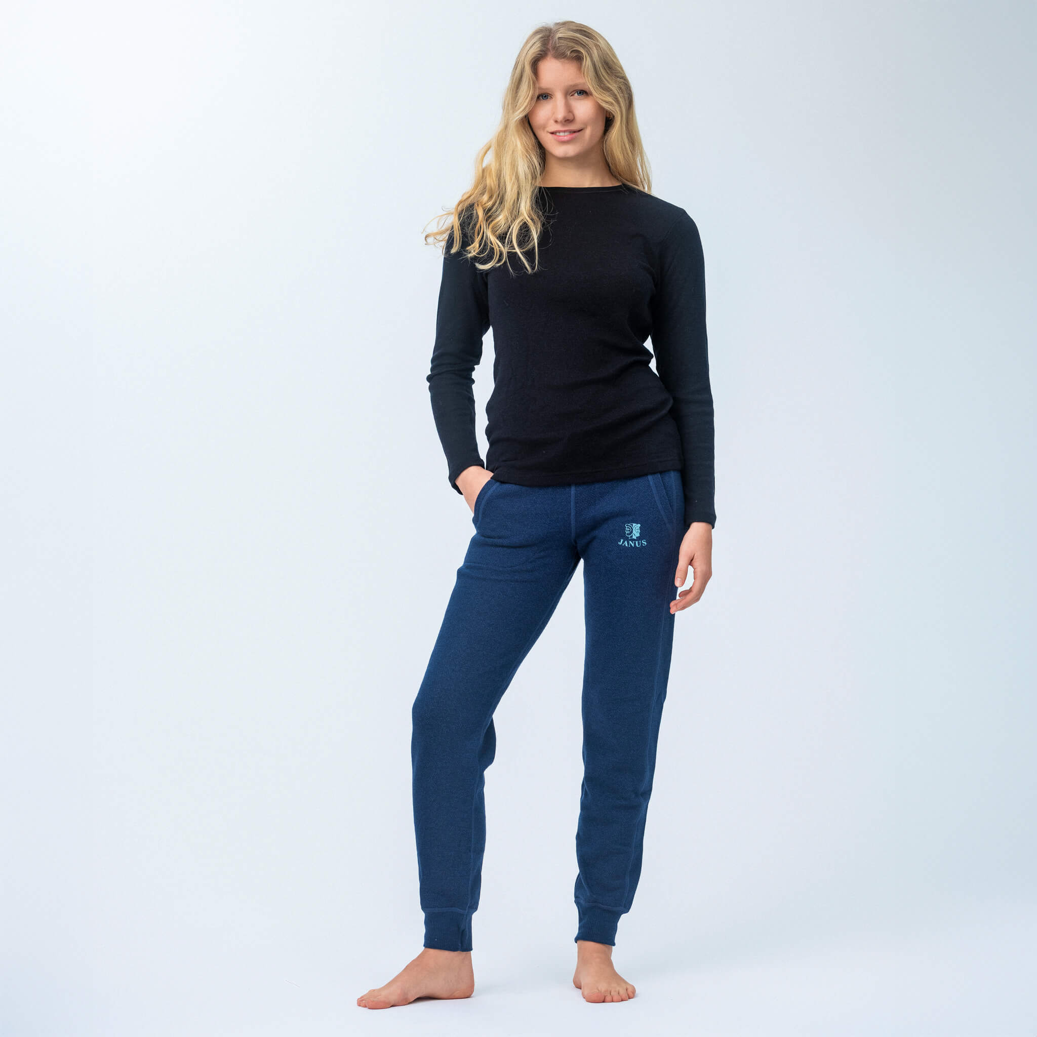 A woman with long blonde hair stands barefoot against a light background, smiling softly with one hand in her pocket. She wears a black long-sleeve shirt and Janusfabrikken Pants merino - Designwool 350 Women in blue.