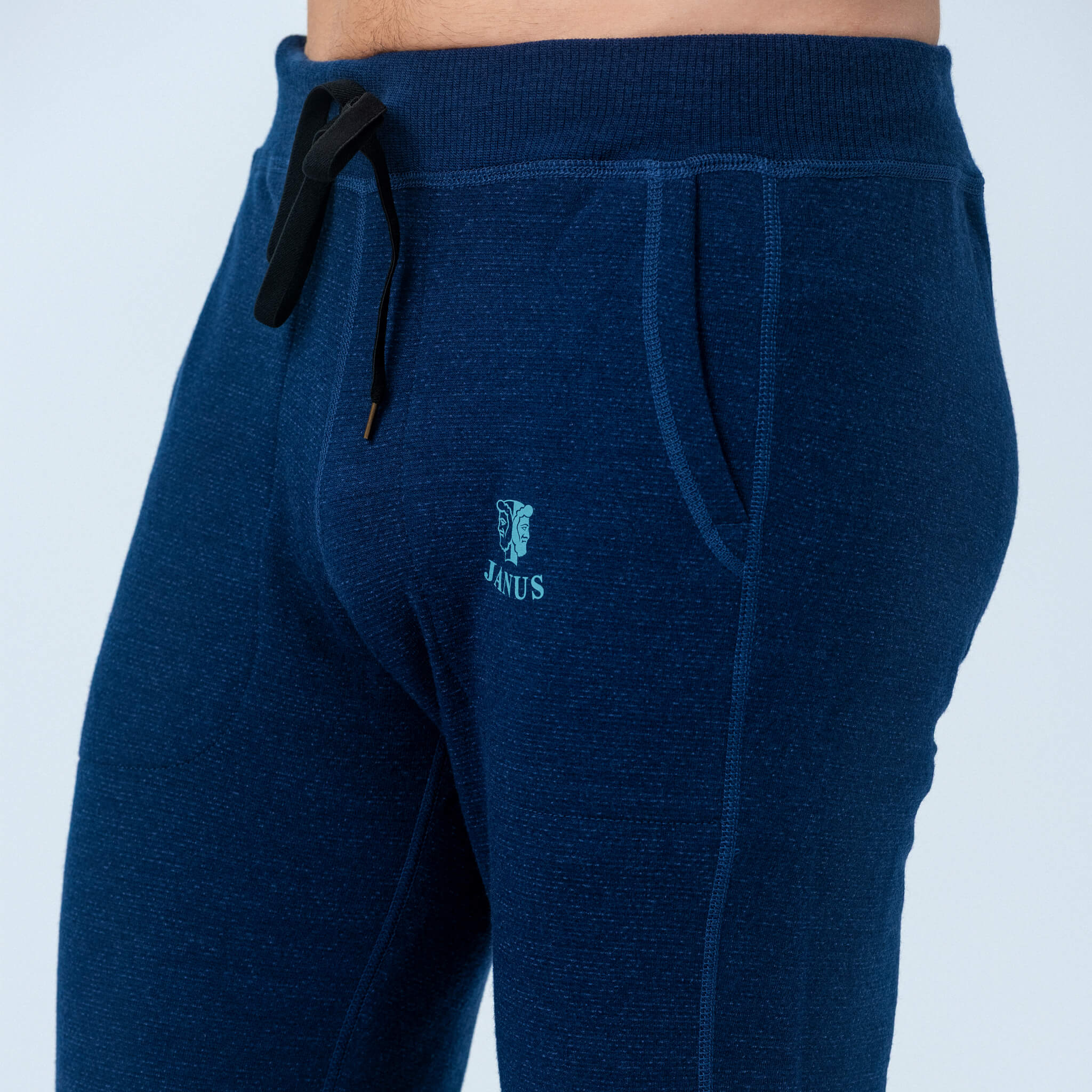 Close-up of a person wearing Pants merino - Designwool 350 Men in dark blue with a black drawstring and the JANUS logo by the pocket, set against a plain light-colored background.