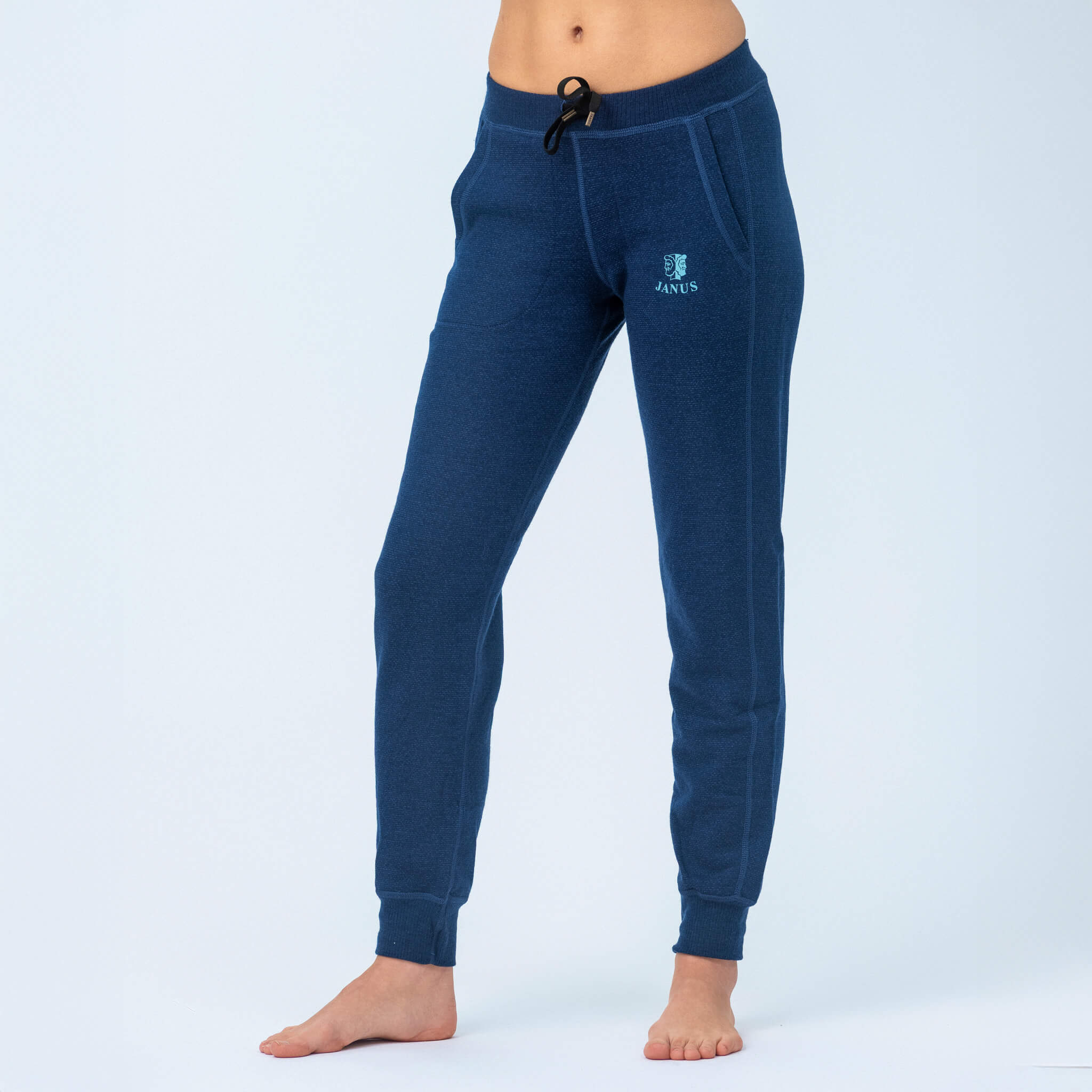 A person wearing blue Janus Wool Pants merino - Designwool 350 Women joggers with pockets, drawstring waistband, and a crest logo on the left thigh stands barefoot against a plain light background.