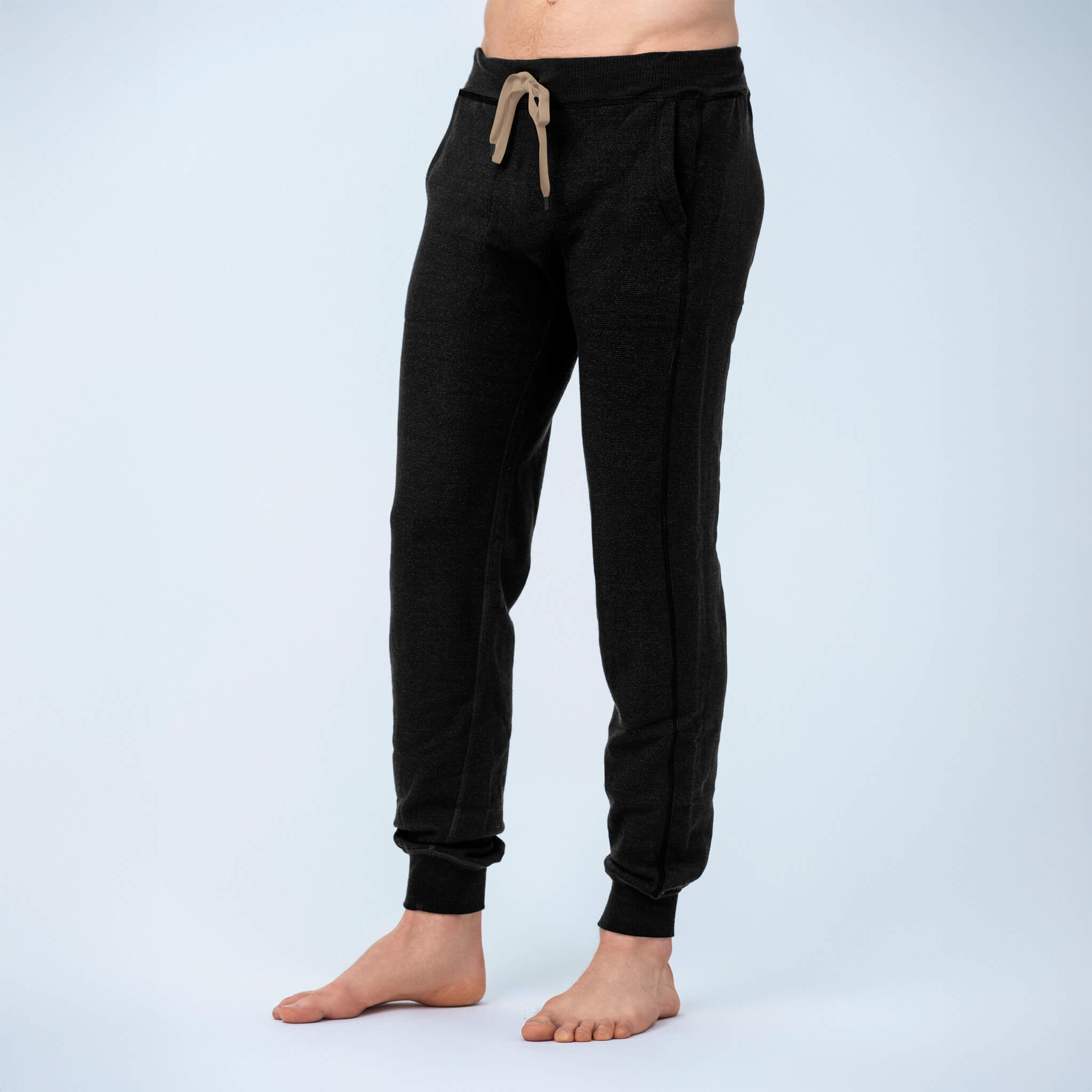 A person stands barefoot in black Pants merino - Designwool 350 Men with an elastic waistband and brown drawstring, against a plain light background.