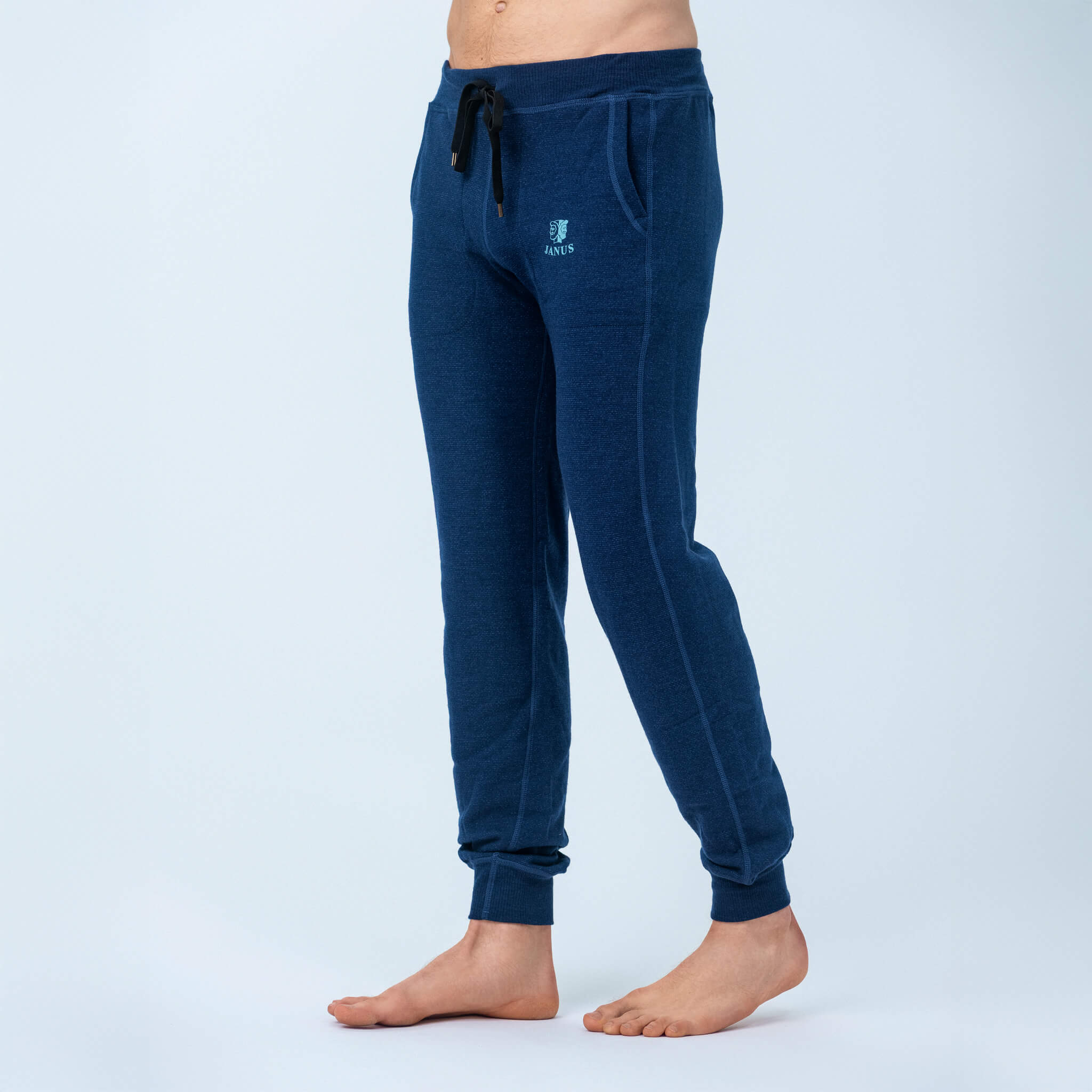 A person stands barefoot against a plain light background wearing Janus Wool Pants merino - Designwool 350 Men in navy blue, featuring pockets and a small logo for a comfortable, casual look.