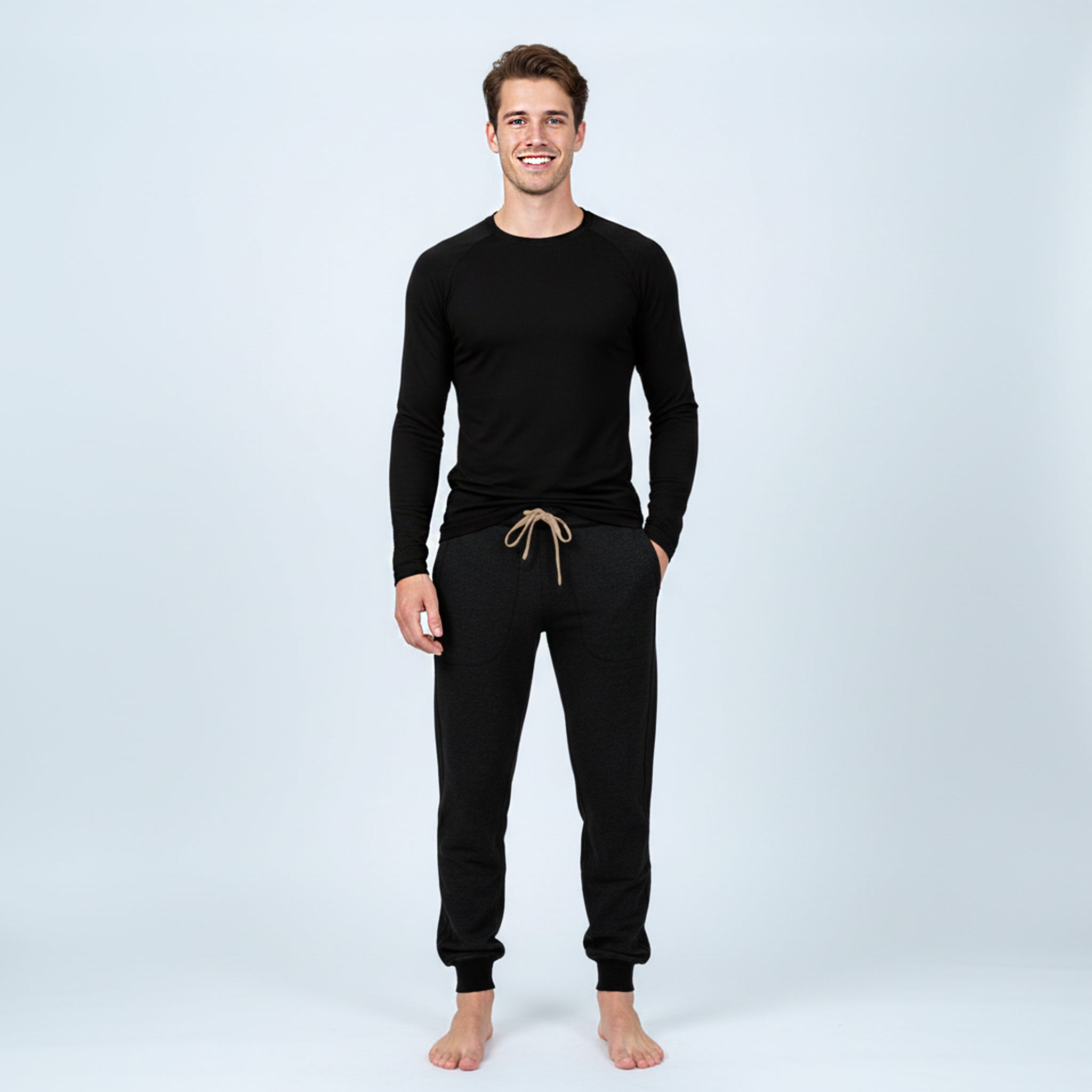 A man stands barefoot against a plain light background, smiling. He wears a long-sleeve black shirt and dark Pants merino - Designwool 350 Men jogger sweatpants with a tan drawstring.