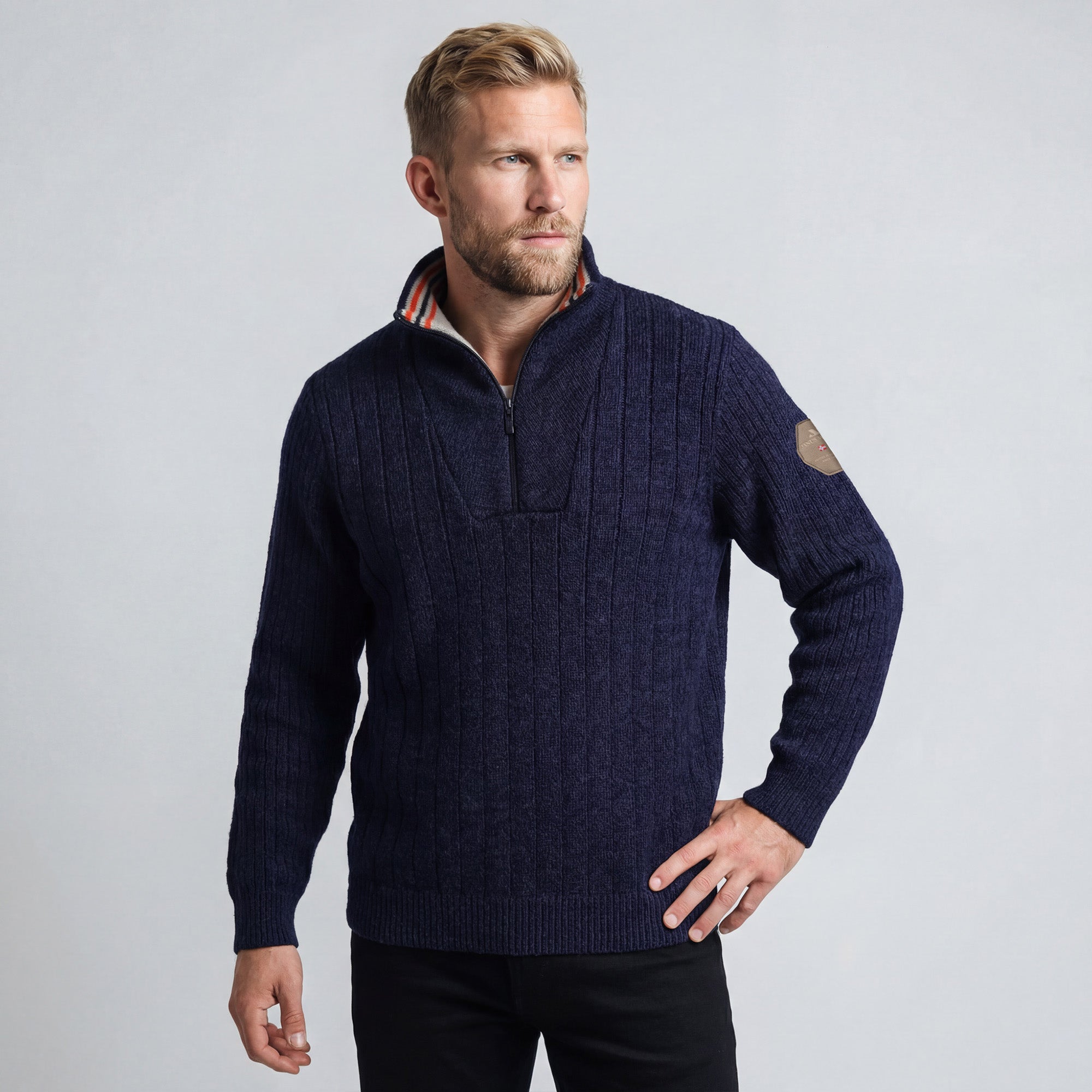 A man with short blond hair and a beard wears the Finse sweater in navy, featuring a half-zip collar, standing with one hand on his hip against a plain light gray background.