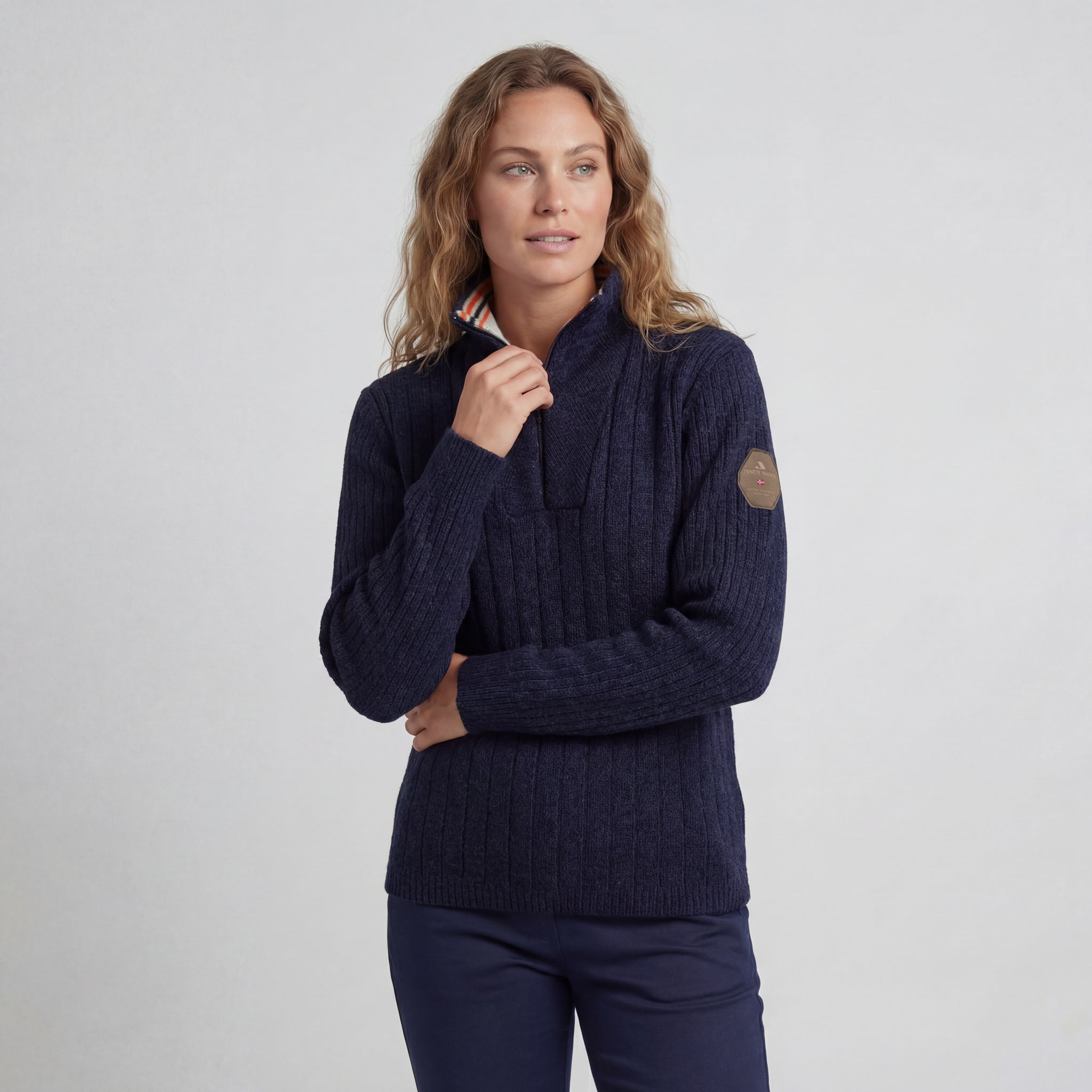 A woman with wavy blonde hair wears the Finse sweater - Navy and dark pants. She stands against a plain light background, looking to the side with a thoughtful expression while touching her chin.