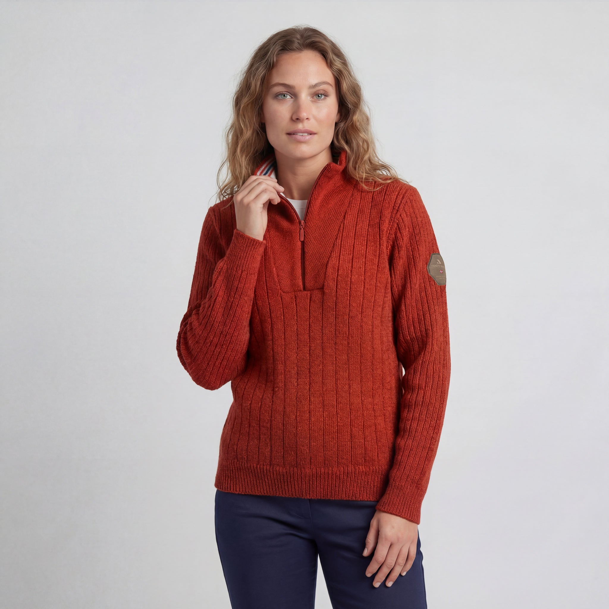 A woman with wavy, light brown hair wears the Finse sweater - Rust, featuring a ribbed knit and half-zip collar, paired with navy pants. She stands against a plain light background.