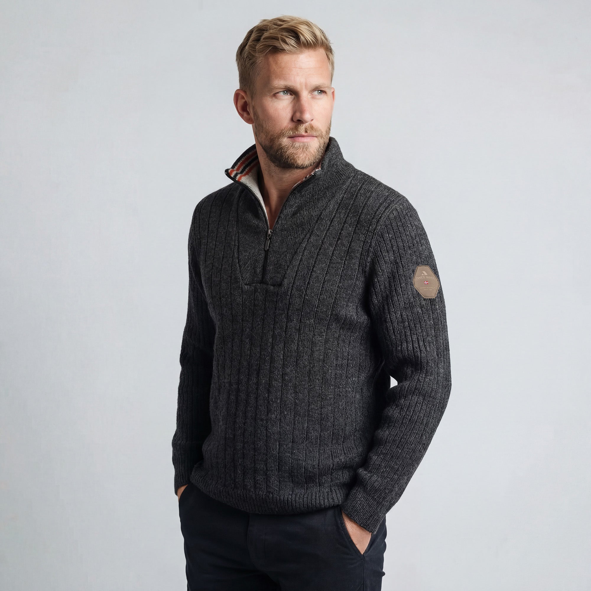 A blond man with a beard wears the Finse sweater - Charcoal, featuring a zip-neck and sleeve patch, paired with black pants. He stands with one hand in his pocket against a light gray background.