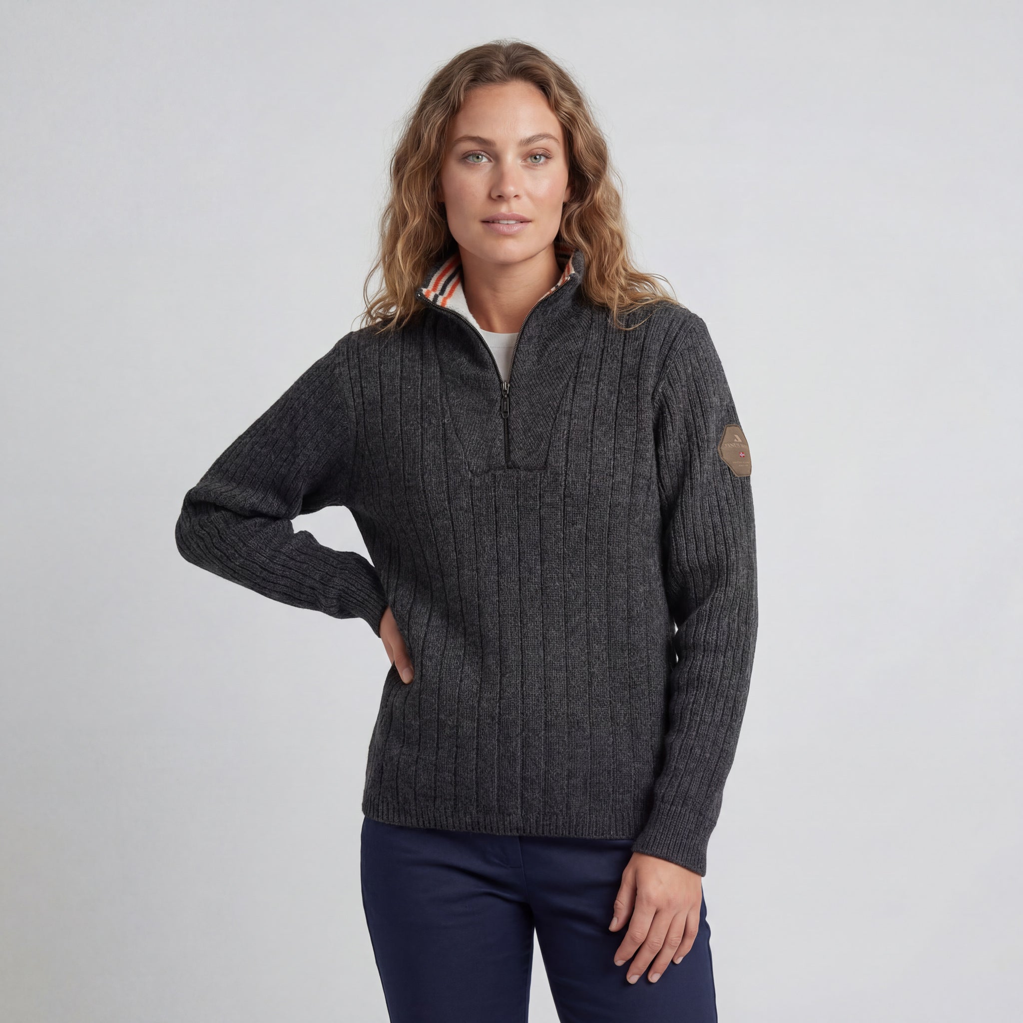 A woman with wavy light brown hair wears the Finse sweater in Charcoal and navy pants, standing with one hand on her hip against a plain light gray background.