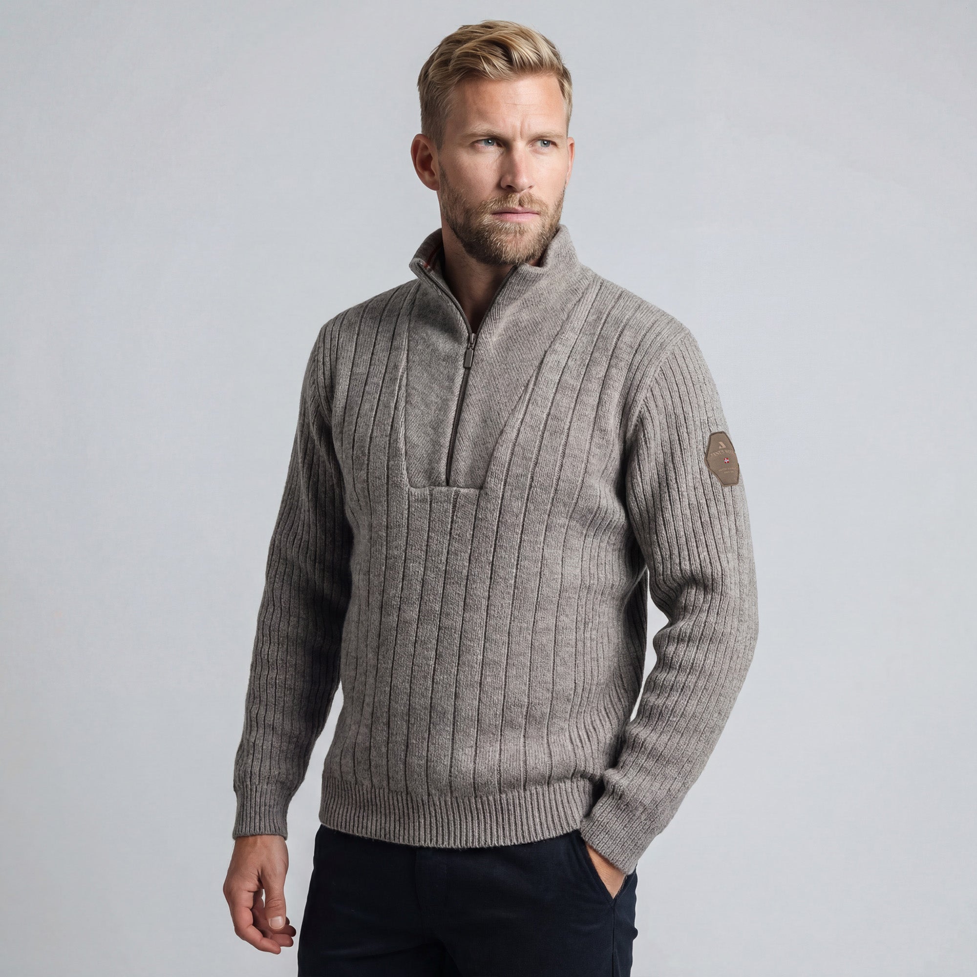 A man with short blond hair and a beard wears the Finse sweater - Greige in greige melange, featuring a ribbed knit, high collar, and half-zip, paired with dark pants against a plain light gray background.