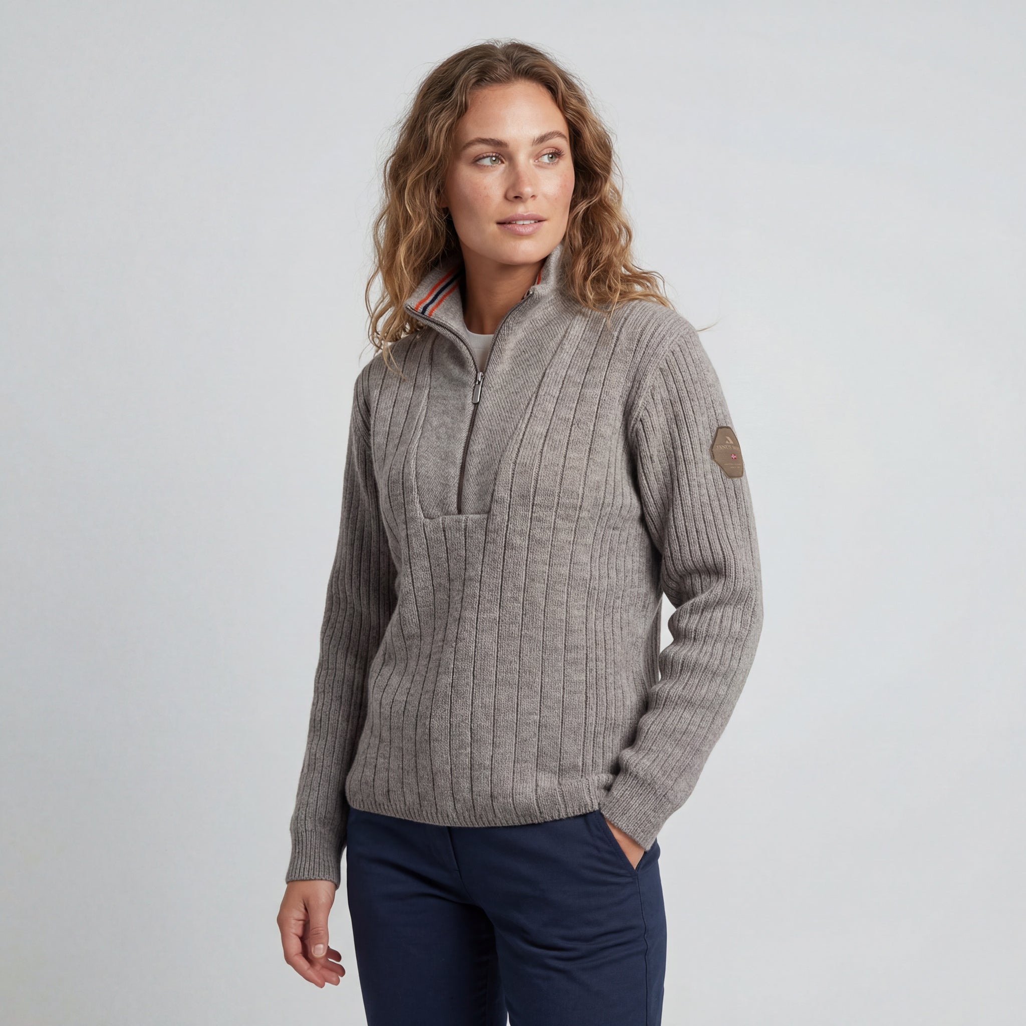 A woman with wavy light brown hair models the Finse sweater - Greige, paired with navy pants, standing against a plain light gray background and looking slightly to her left with a relaxed expression.