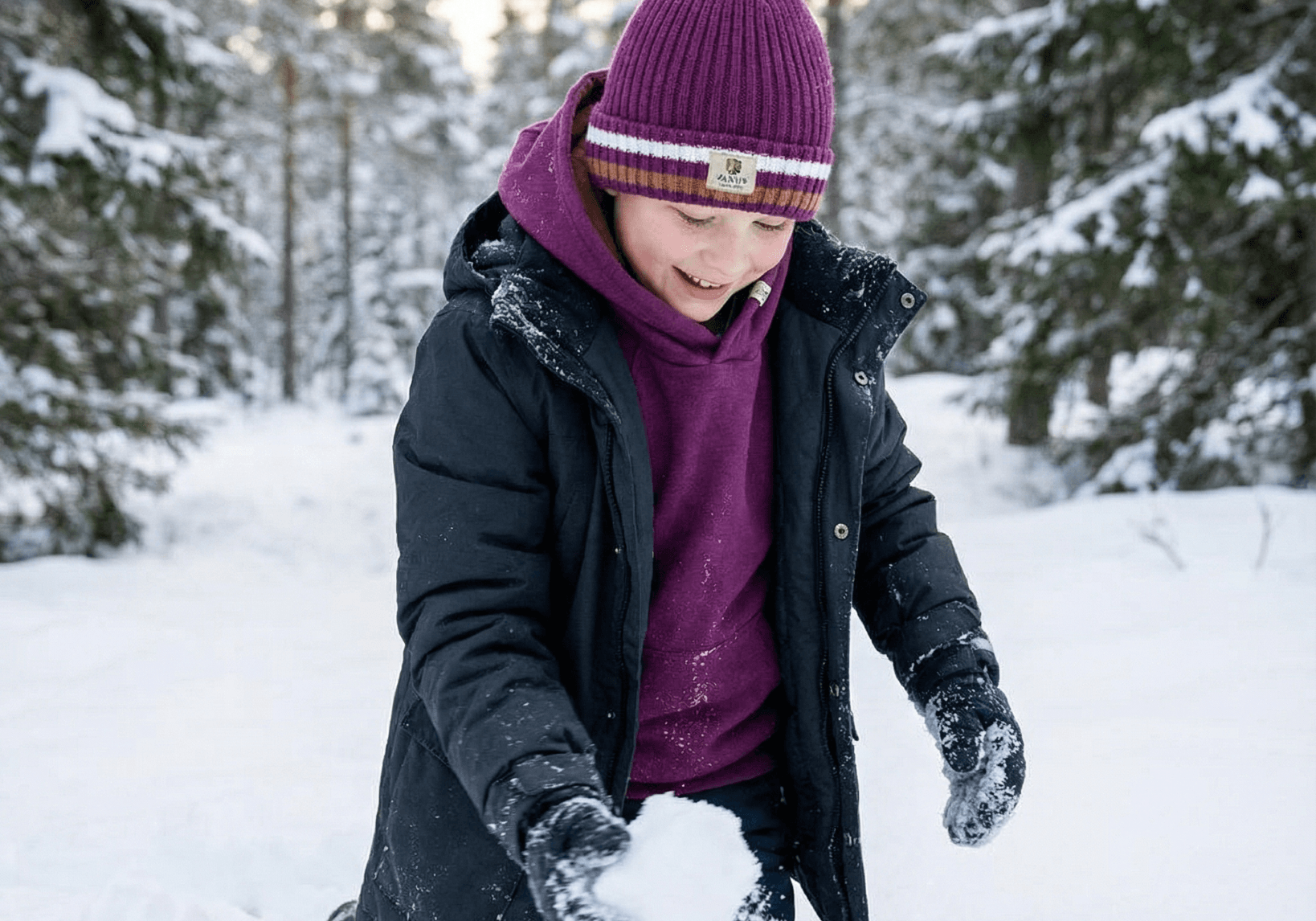 A child in a purple hat and Janus Wool's Polo with zip merino - Janus Go 250 Kids sweater kneels in snowy woods, smiling while making a snowball among the trees.