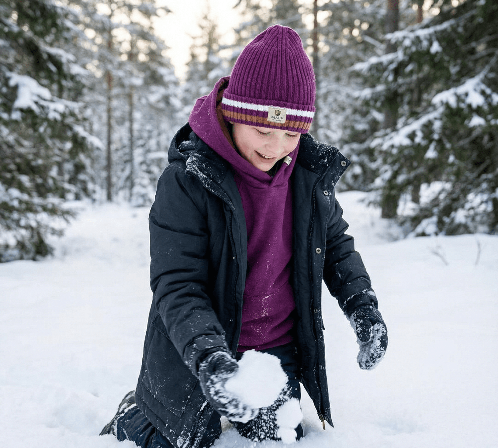 A child in a purple hat and Janus Wool's Polo with zip merino - Janus Go 250 Kids sweater kneels in snowy woods, smiling while making a snowball among the trees.