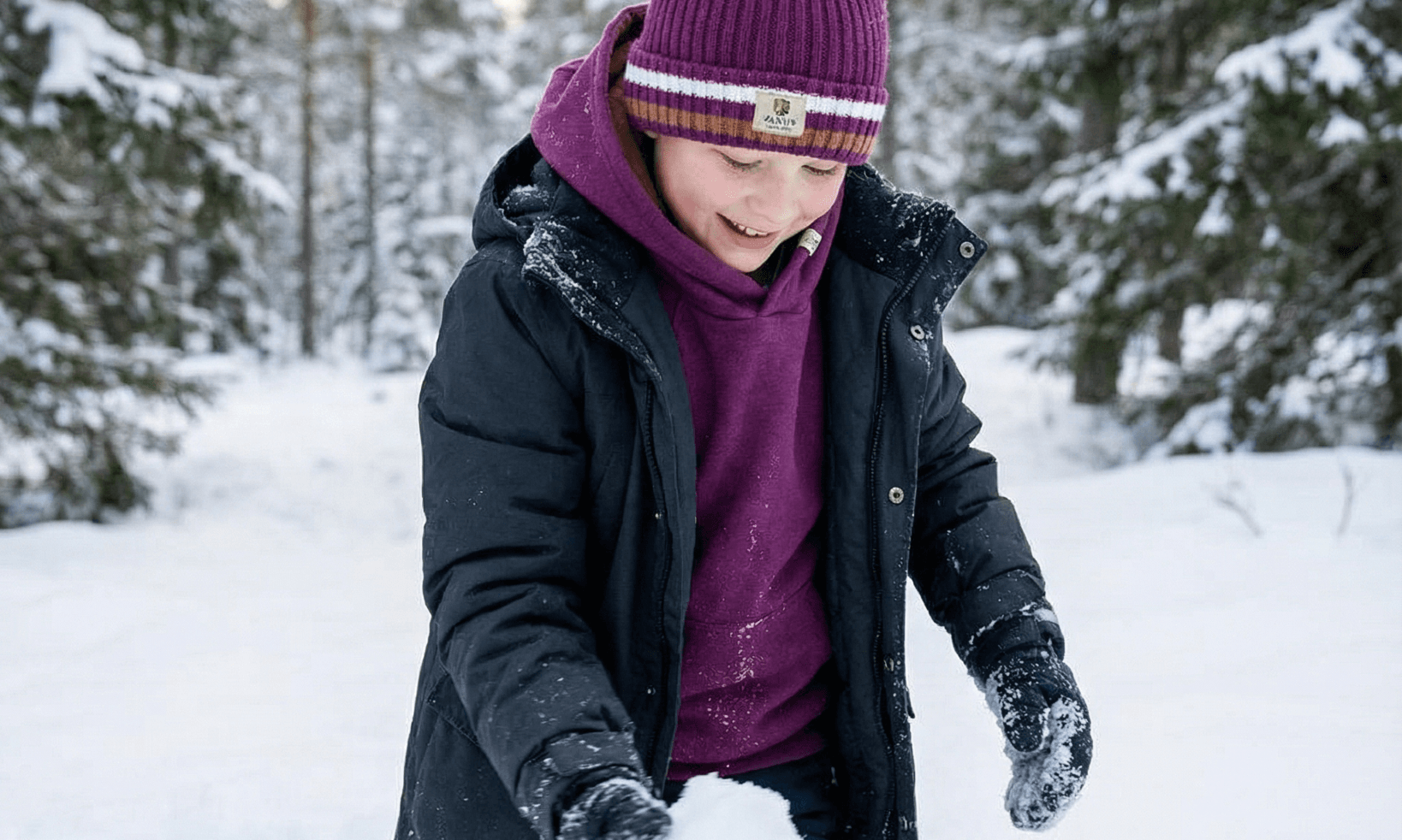 A child in a purple hat and Janus Wool's Polo with zip merino - Janus Go 250 Kids sweater kneels in snowy woods, smiling while making a snowball among the trees.