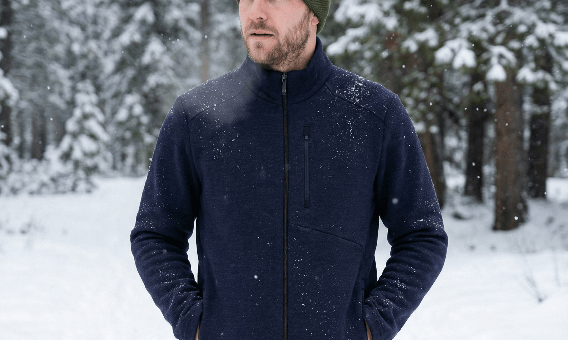 A man wearing a green merino wool beanie and navy jacket stands outdoors in a snowy forest, with trees and fresh snow in the background. He has his hands in his jacket pockets and looks slightly to the side.
