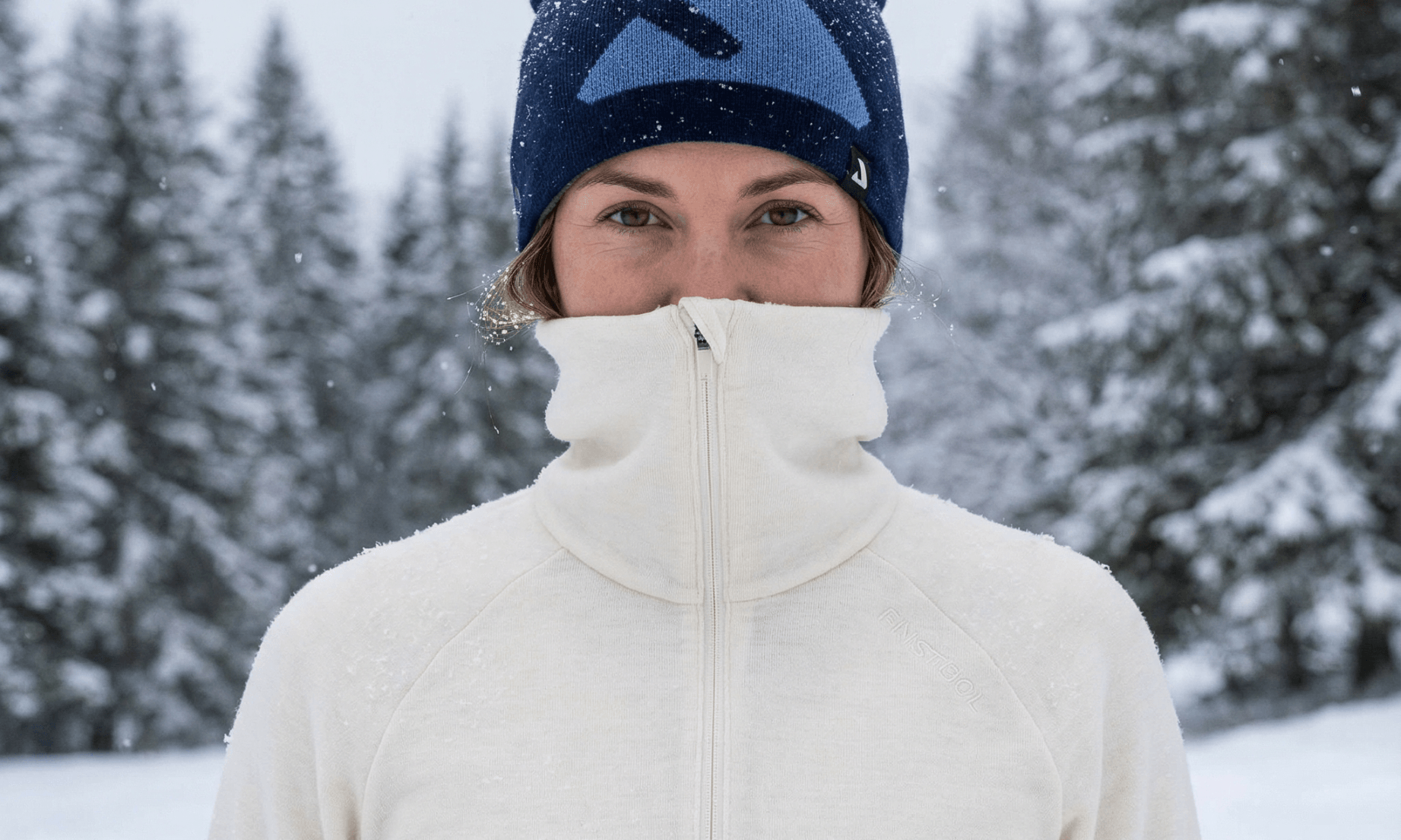 A person in a cream merino wool zip-up jacket and blue winter hat stands outside in falling snow, surrounded by snow-covered trees, with their jacket zipped up to their nose.