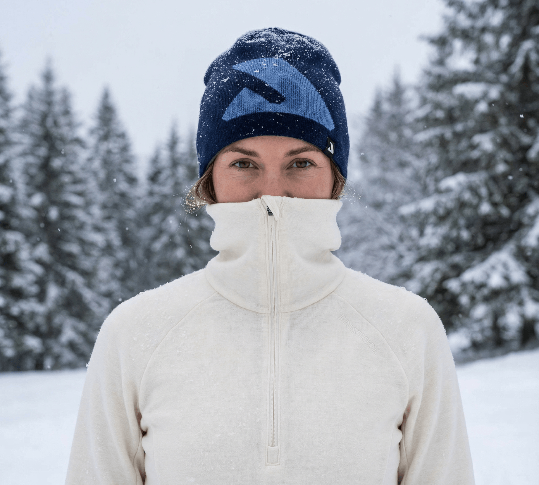A person in a cream merino wool zip-up jacket and blue winter hat stands outside in falling snow, surrounded by snow-covered trees, with their jacket zipped up to their nose.