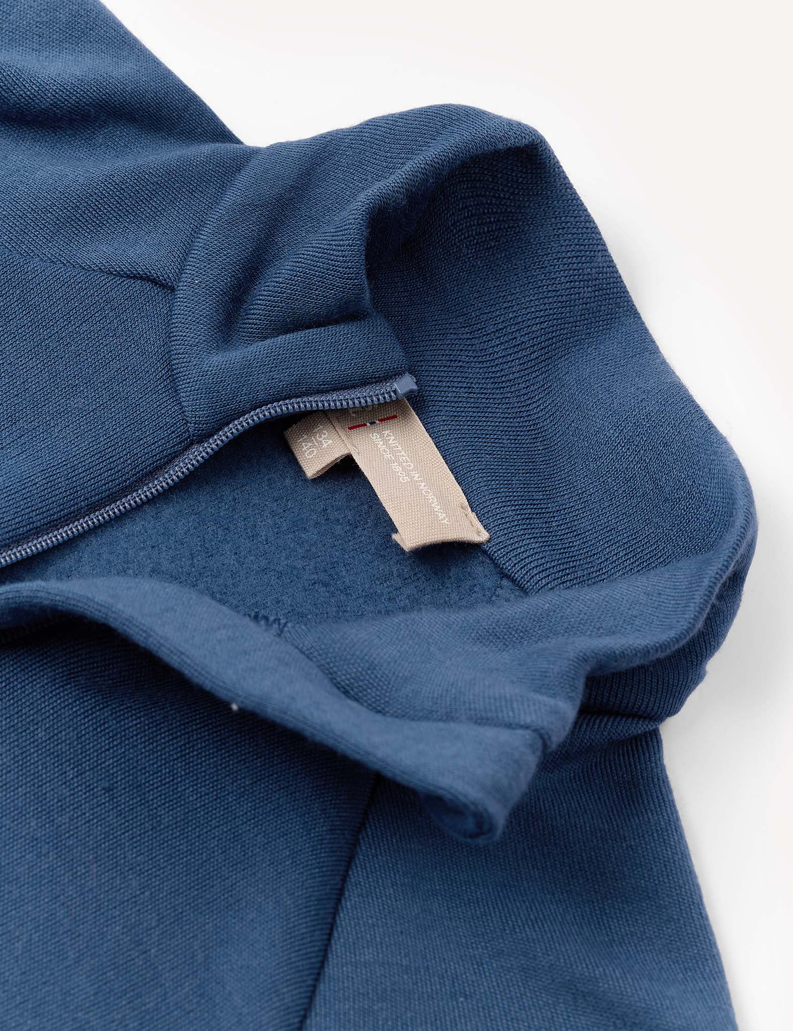 A close-up of a blue Half Zip – Merino Kids AirWool shirt highlights the hood, zipper, and a beige fabric clothing tag sewn into the neckline.