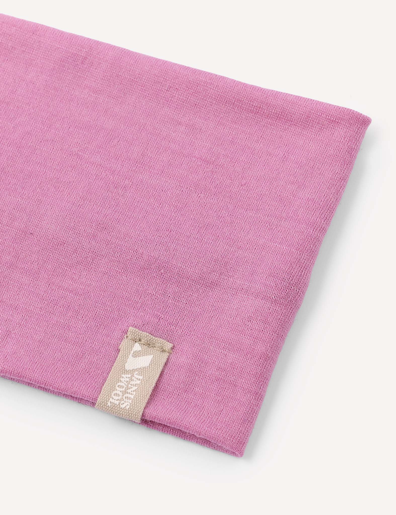 A close-up of the Headband – Merino Kids Light shows a folded pink merino wool headband with a beige tag sewn on, featuring a small white logo and text. The background is plain and light-colored.