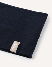 A close-up of the “Headband – Merino Kids Light,” folded in dark navy merino wool, featuring a beige tag with white text and a small heart symbol. The soft fabric is displayed against a light background.