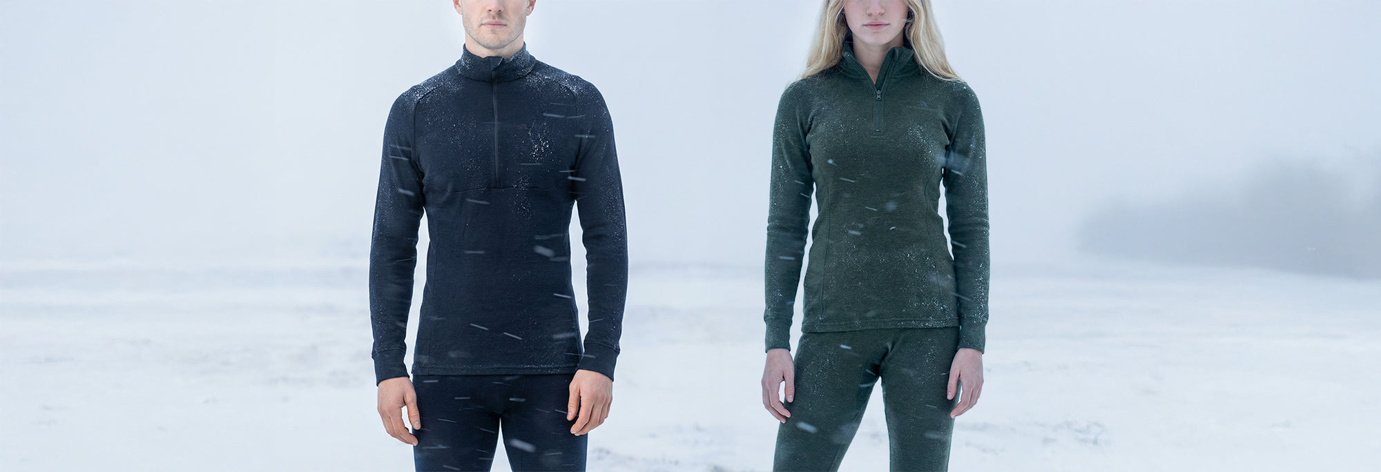 A man and a woman stand side by side in snowy, windy weather, wearing warm, long-sleeve merino wool zip-up tops and matching pants. The background is a foggy, snow-covered landscape.