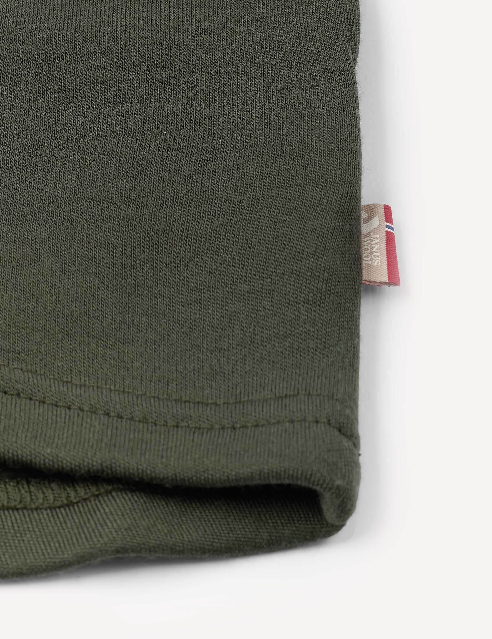 Close-up of the hem of the green Jacket – Merino Kids AirWool, showing a small fabric tag with red, white, and blue stripes sewn into the side seam.
