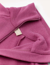 A close-up of the Jacket – Merino Kids AirWool in a mauve or pinkish-purple shade highlights its soft brushed AirWool fabric texture, with an off-white clothing tag partially visible near the seam.