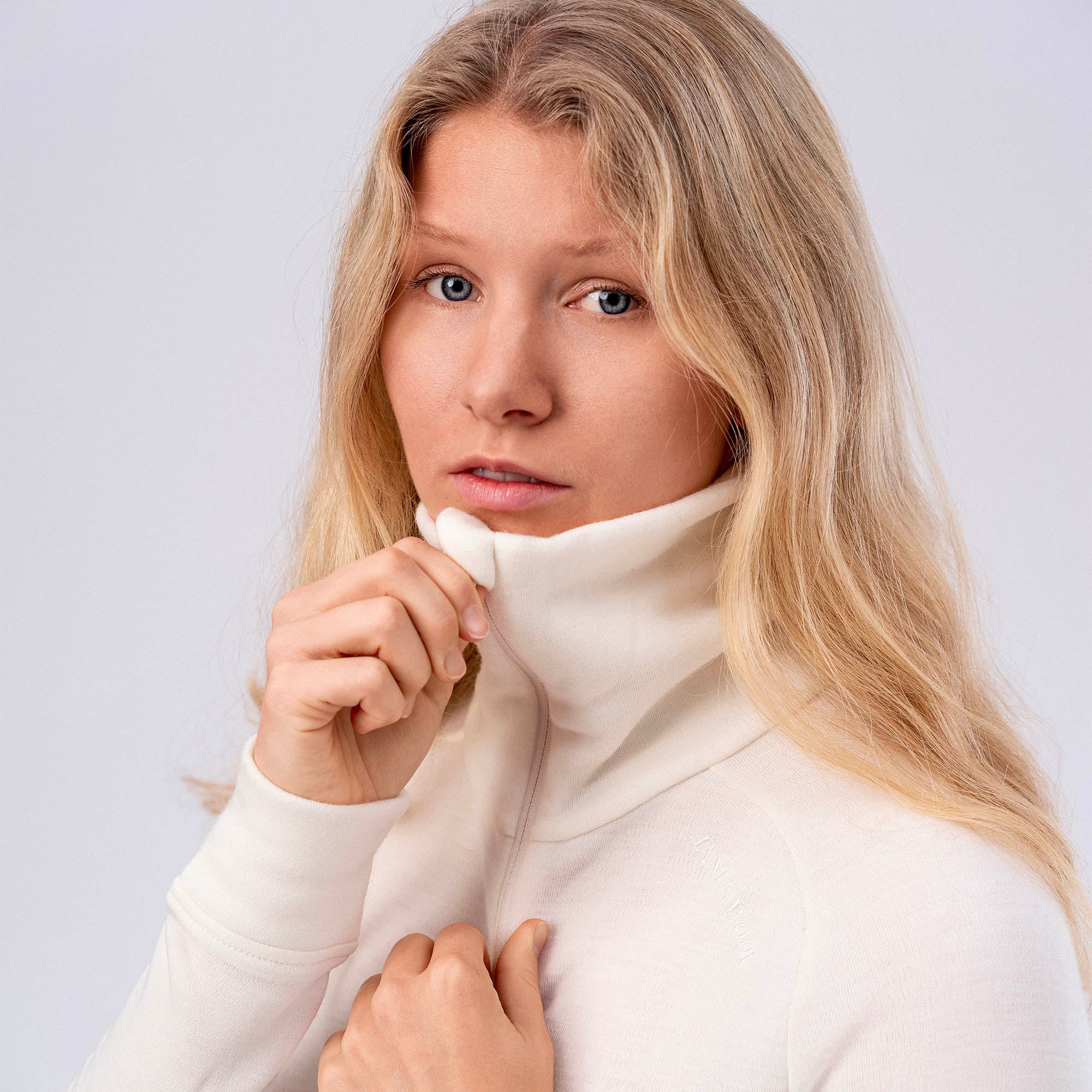 A woman with long blonde hair and blue eyes models the Polo Zip Merino – AirWool 260 Women, a white, breathable wool turtleneck, gently adjusting the collar and looking at the camera against a neutral background.