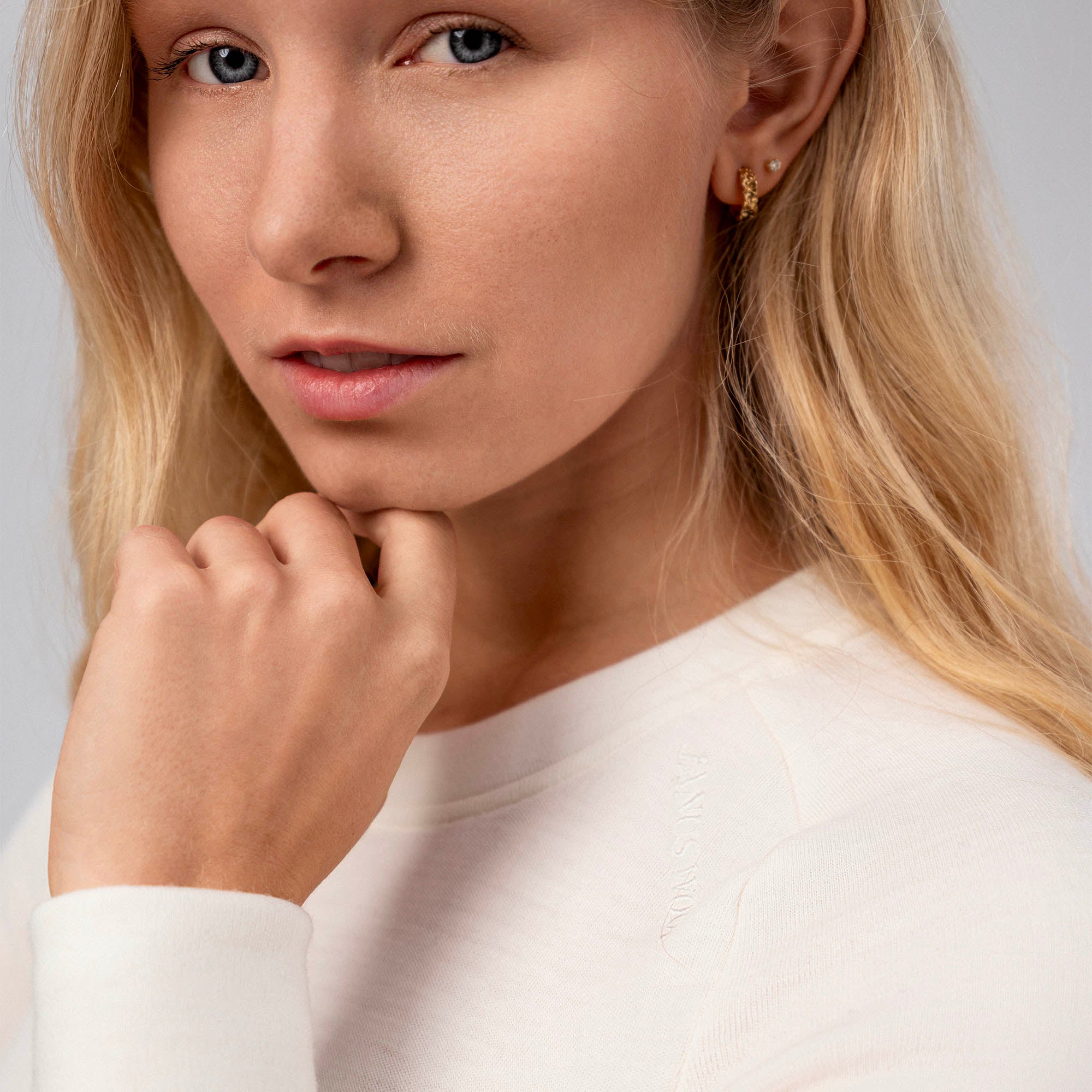 A woman with long blonde hair wears the Jersey Merino – AirWool 260 Women, a white breathable top, and small gold earrings. She gazes at the camera with her hand thoughtfully near her chin against a light gray background.
