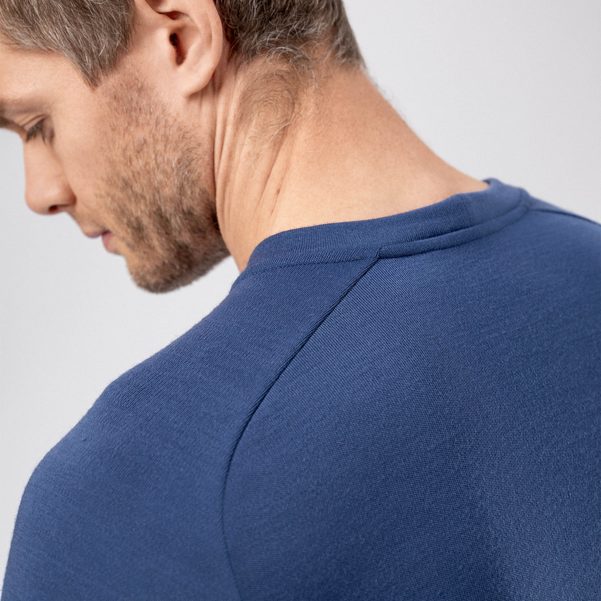 Close-up of a man’s neck and shoulder in the Jersey Merino – AirWool 260 Men, blue crew-neck, facing left with head slightly down. The image highlights the shirt’s texture, seam detail, warmth, and breathability against a plain light background.