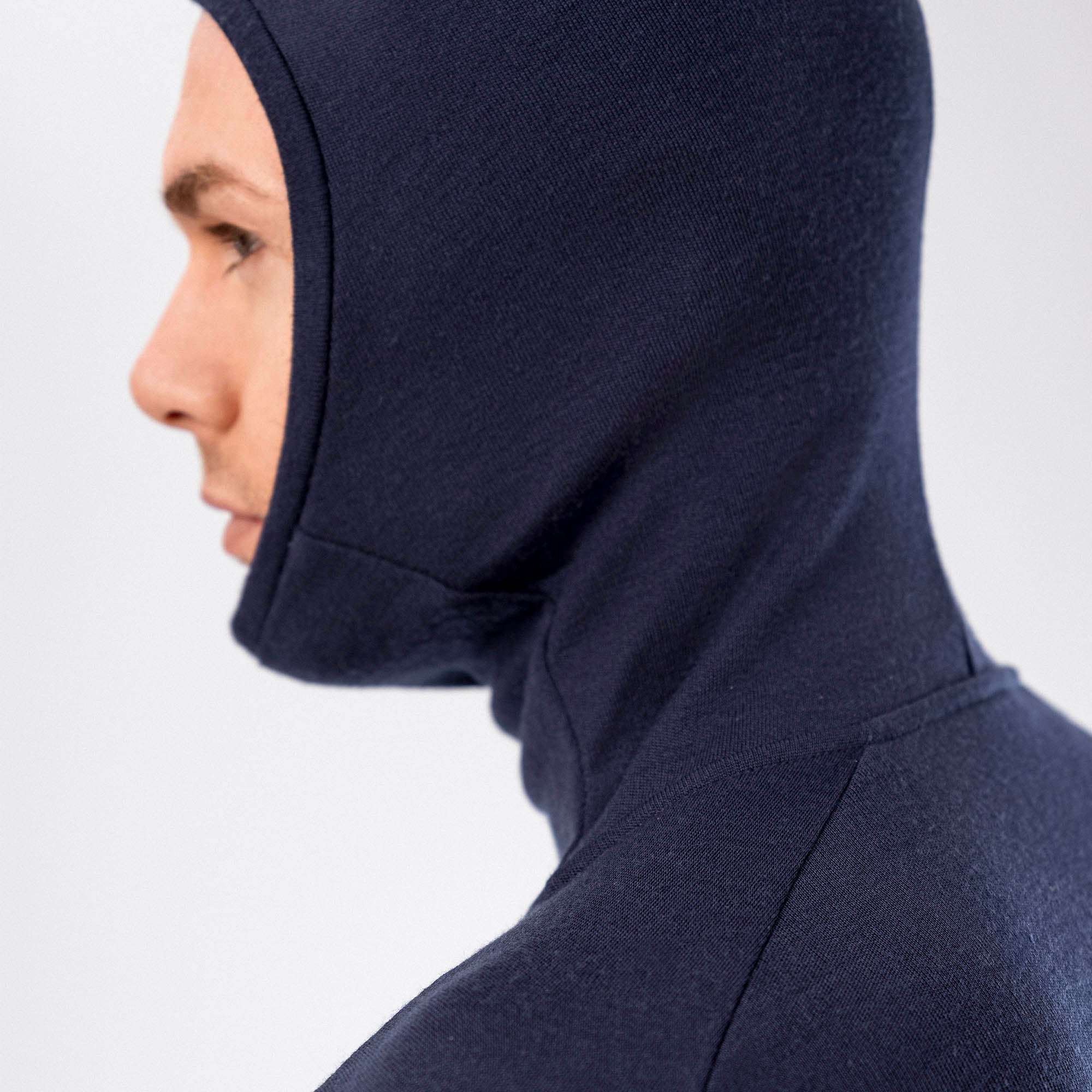 A person in profile wears the Hooded Jersey Merino – AirWool 260 Men, a fitted dark blue merino wool hooded jersey covering most of their head and neck, set against a plain light background.