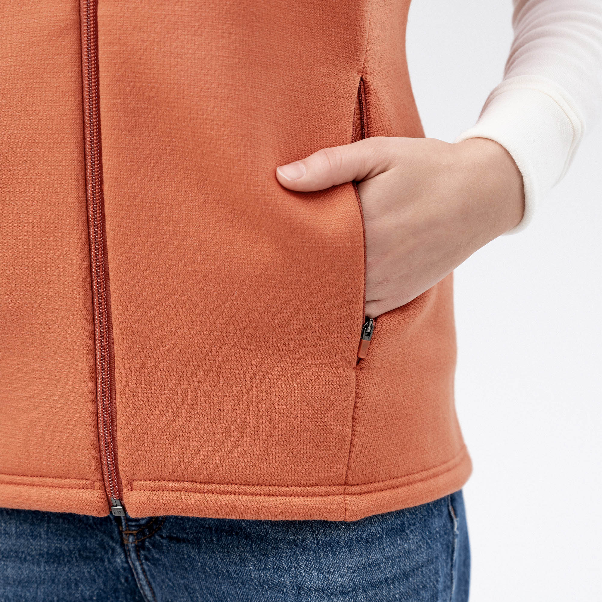 A person in a white long-sleeve shirt, blue jeans, and the Vest Merino – AirWool 350 Women for added warmth has their right hand in the vest pocket.