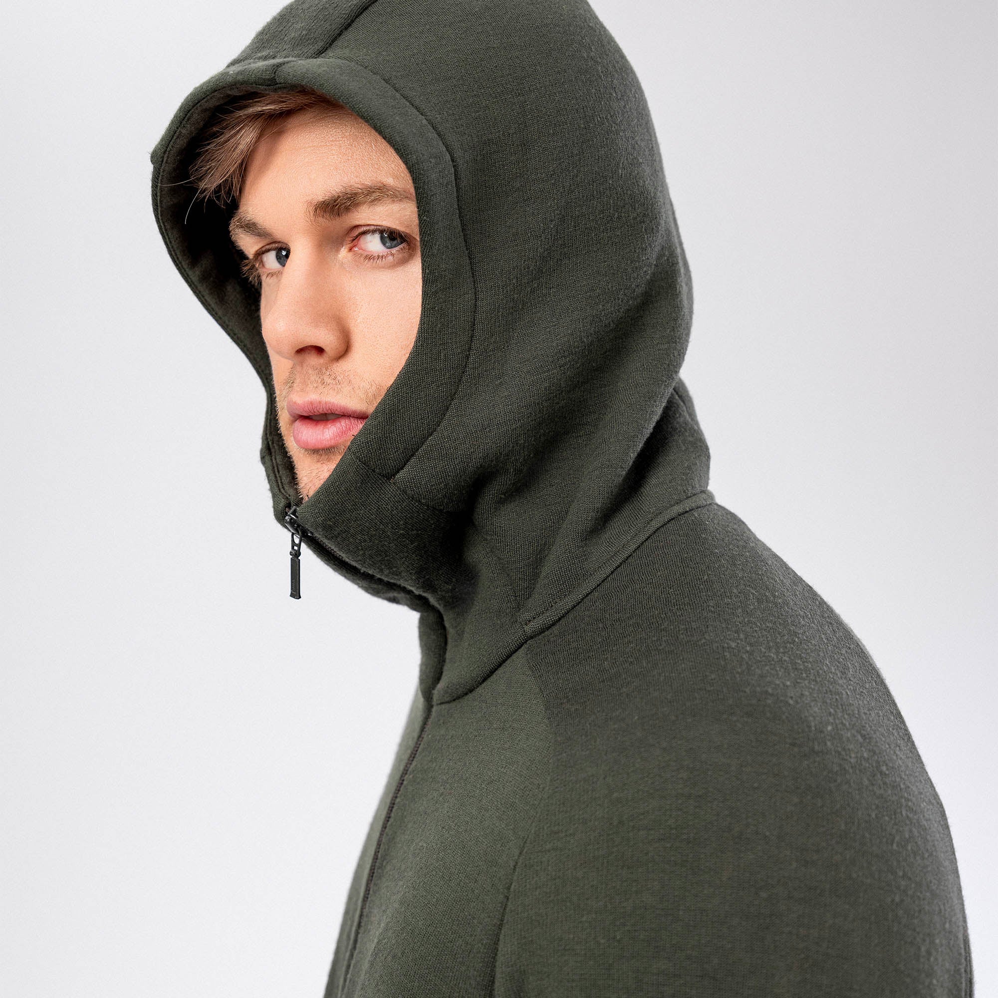 A young man wears the Hooded Jacket Merino – AirWool 280 Men in dark green with the hood up, looking at the camera with a neutral expression against a plain light background.