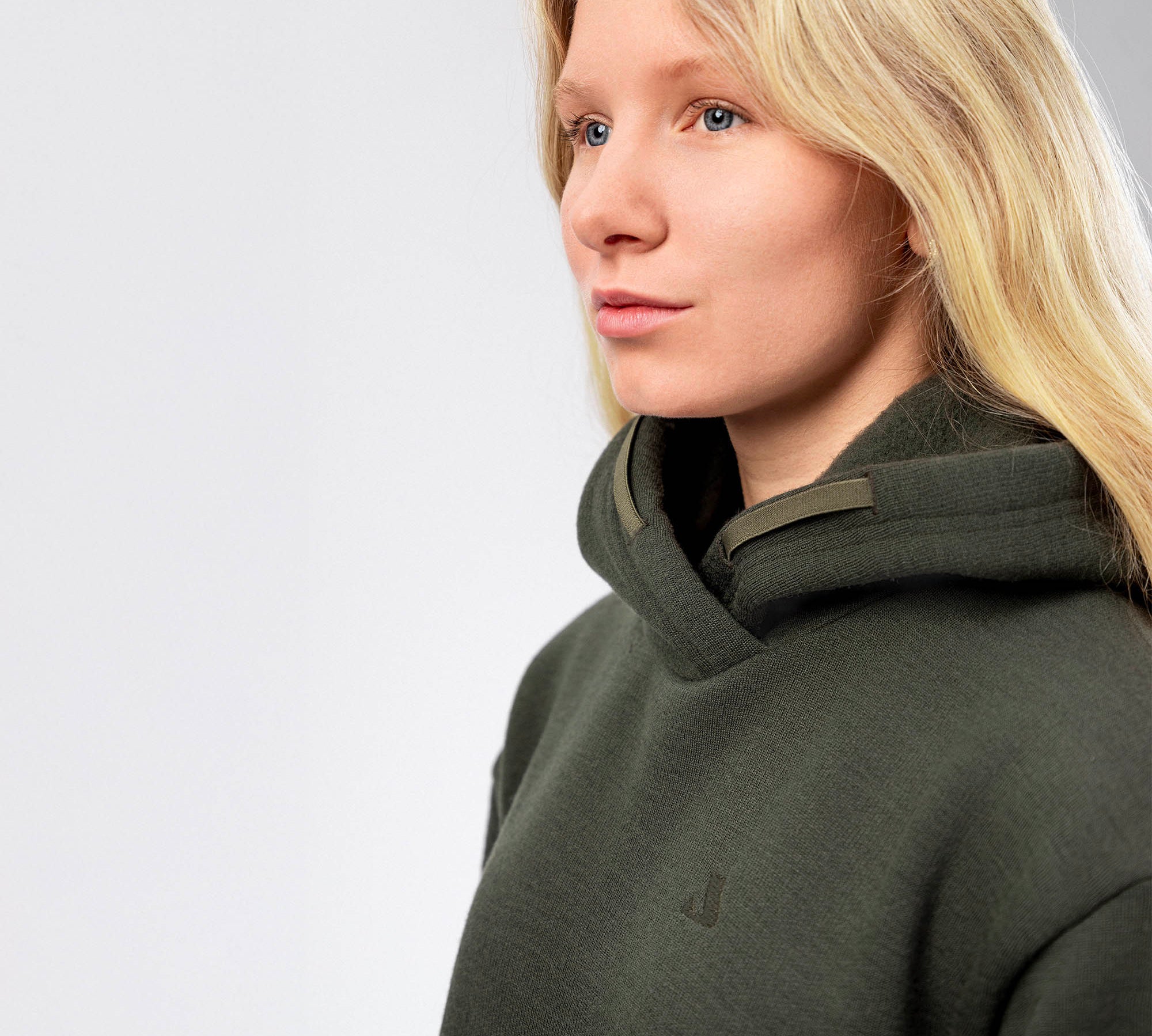 A woman with long blonde hair, wearing the Hoodie Merino – AirWool 280 Women in dark green, gazes slightly to the side against a light gray background.
