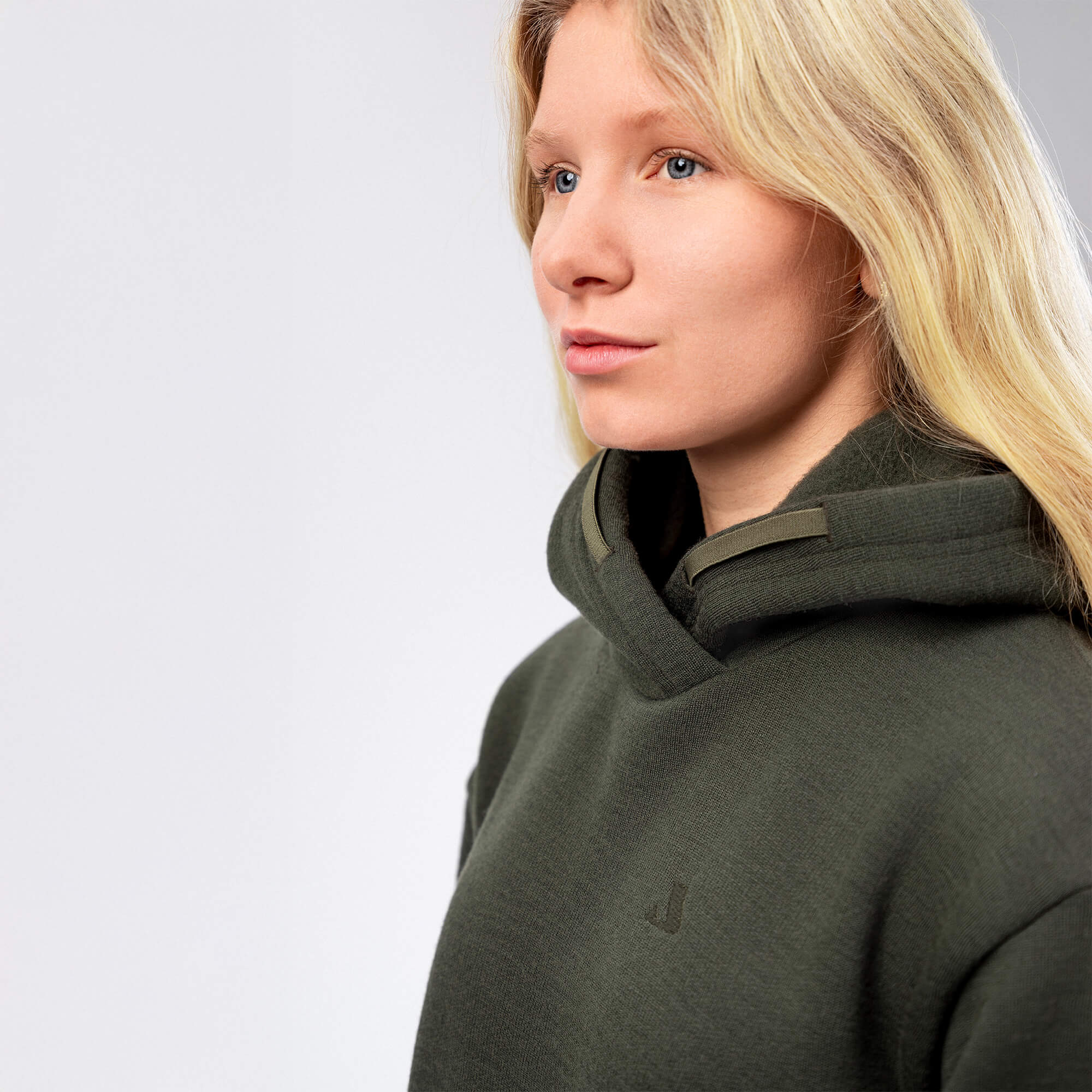 A woman with long blonde hair wearing an olive green merino wool hoodie looks ahead with a neutral expression against a plain light background.
