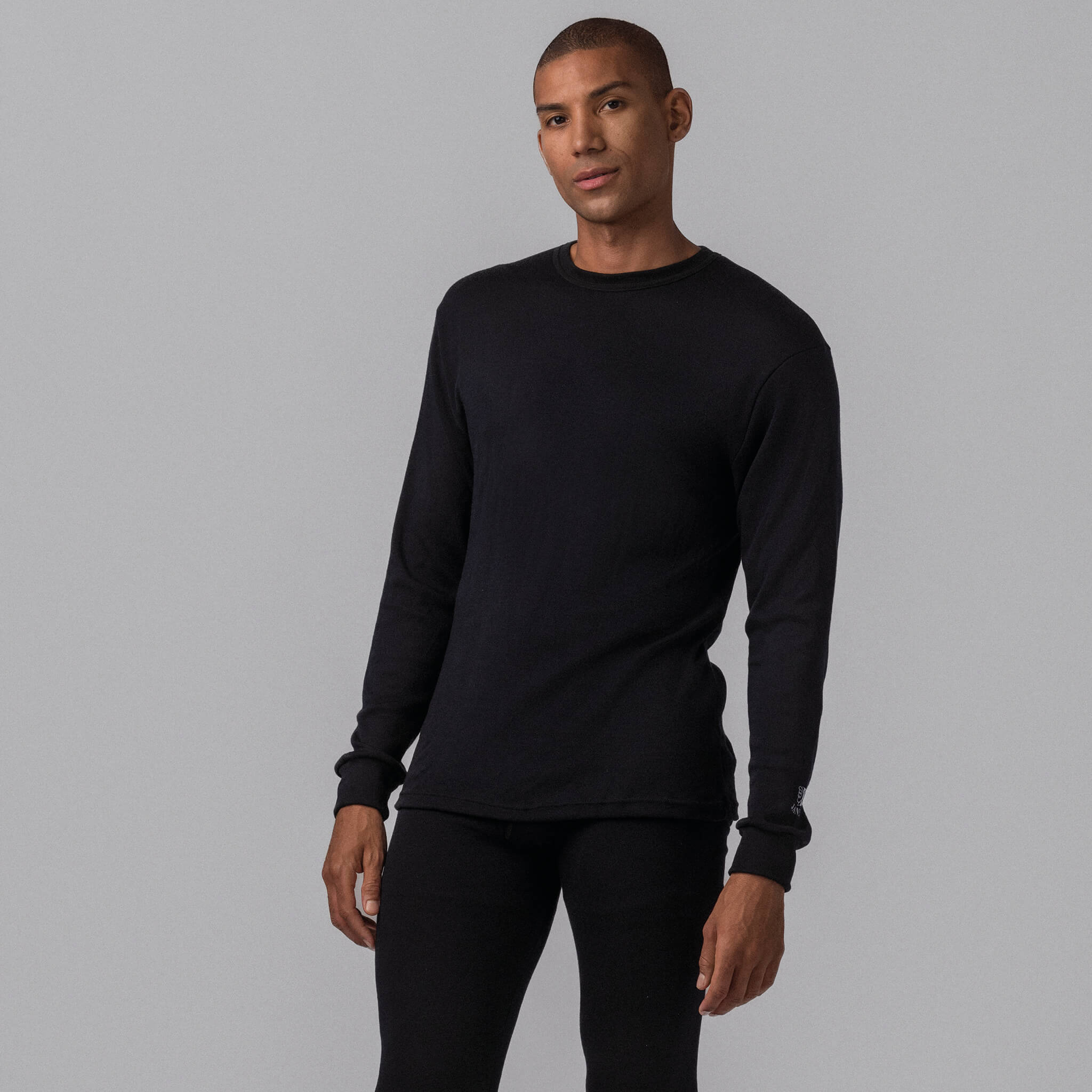 A person stands against a plain gray background, confidently wearing the Janus Jersey merino - Black Wool 200 Men long-sleeve shirt and matching black pants, looking directly at the camera.