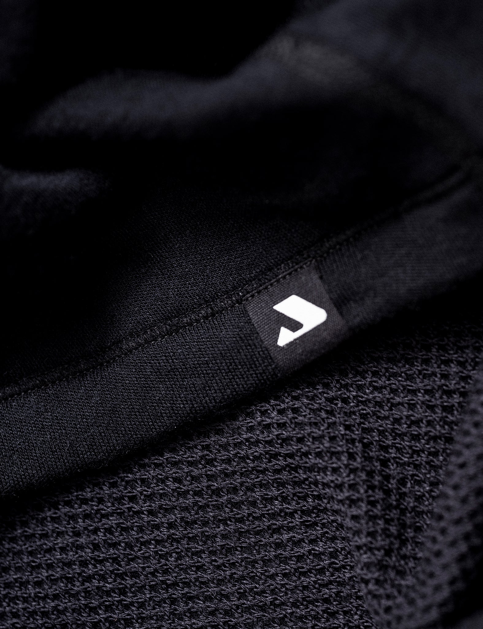 Close-up of the Norse Merino Unisex Double Layer Jacket in black showcasing its textured yet smooth fabric, with a small black tag featuring a white geometric logo.