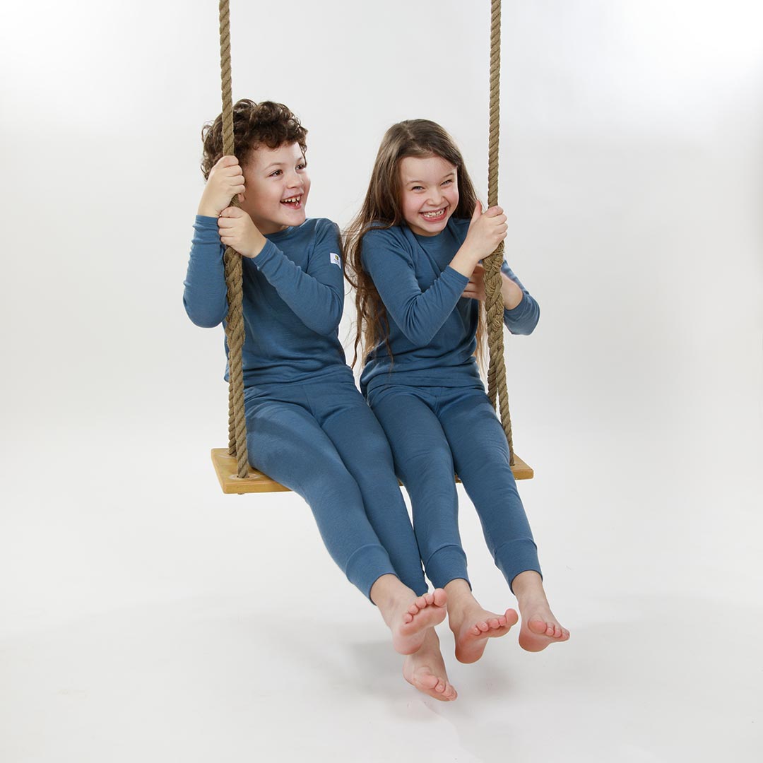 Two children in matching Janus merino blue outfits sitting on a swing against a white background