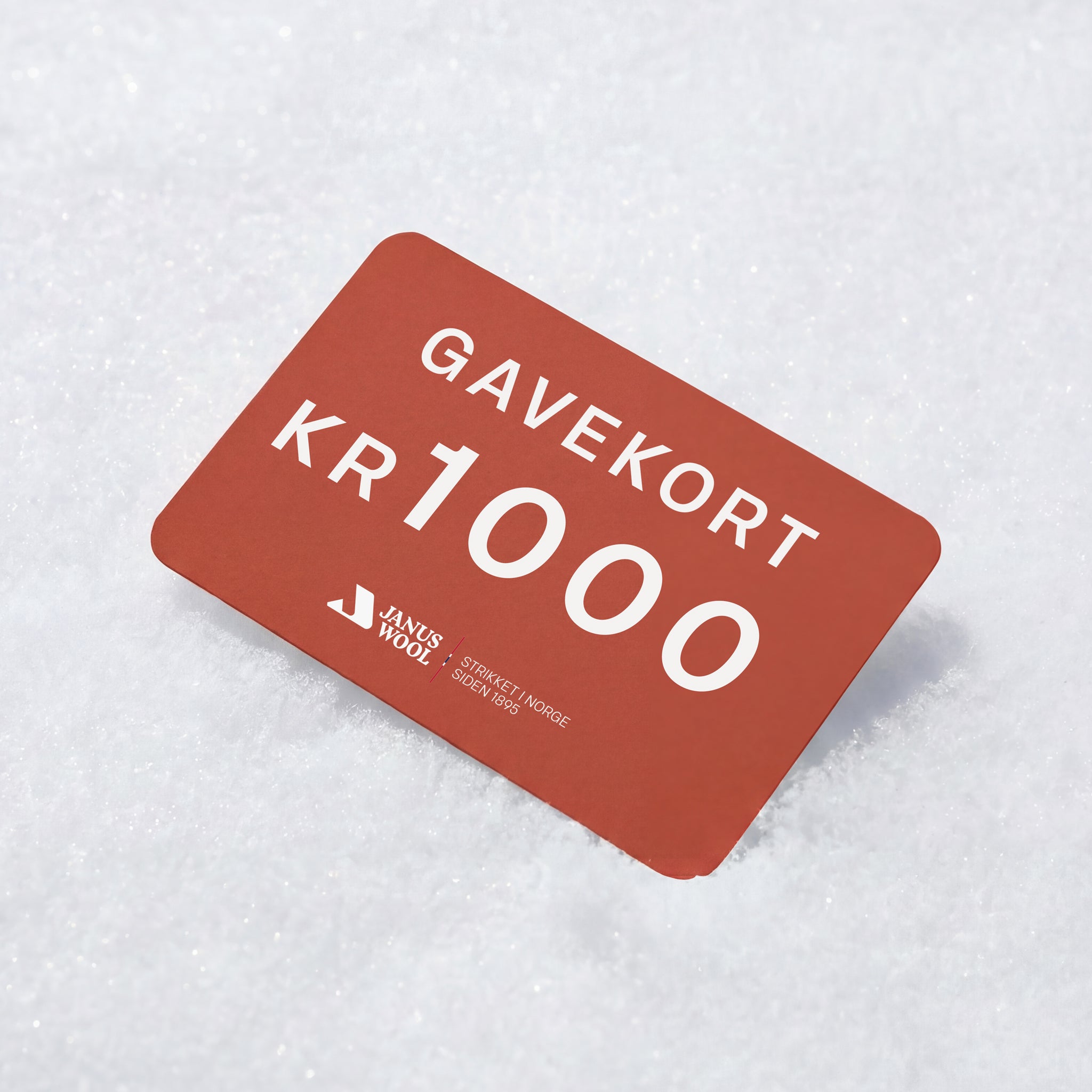 A red Janus Wool Gift Card with white text "Gavekort KR 1000" lies on snow, featuring the Janus Wool brand known for quality merino wool products.