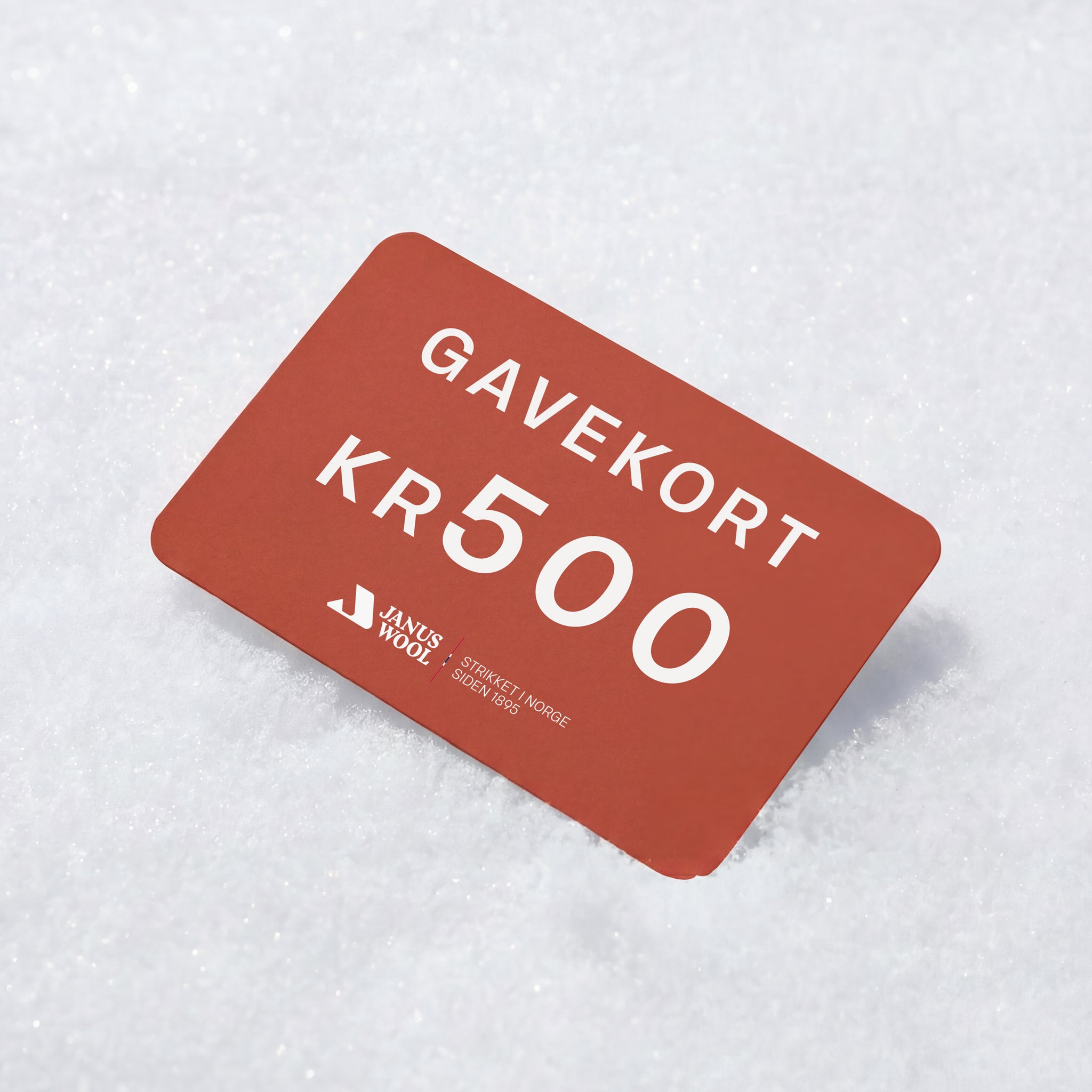 A red Janus Wool Gift Card, with white text displaying GAVEKORT KR 500, rests on a snow-covered surface.