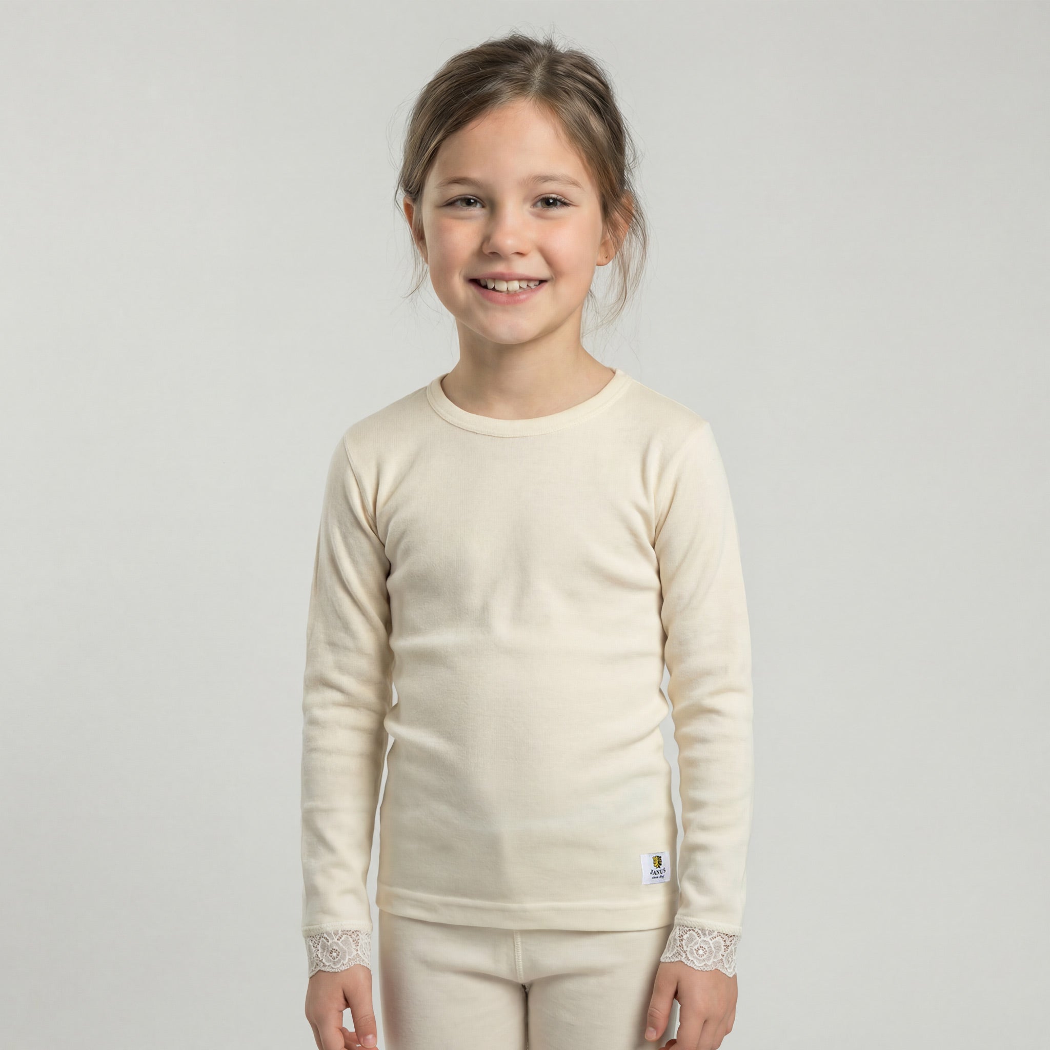 A young girl with brown hair in a ponytail smiles while wearing the "Jersey with laces merino - Janusull 180 Kids" and matching pants, standing against a plain light background.