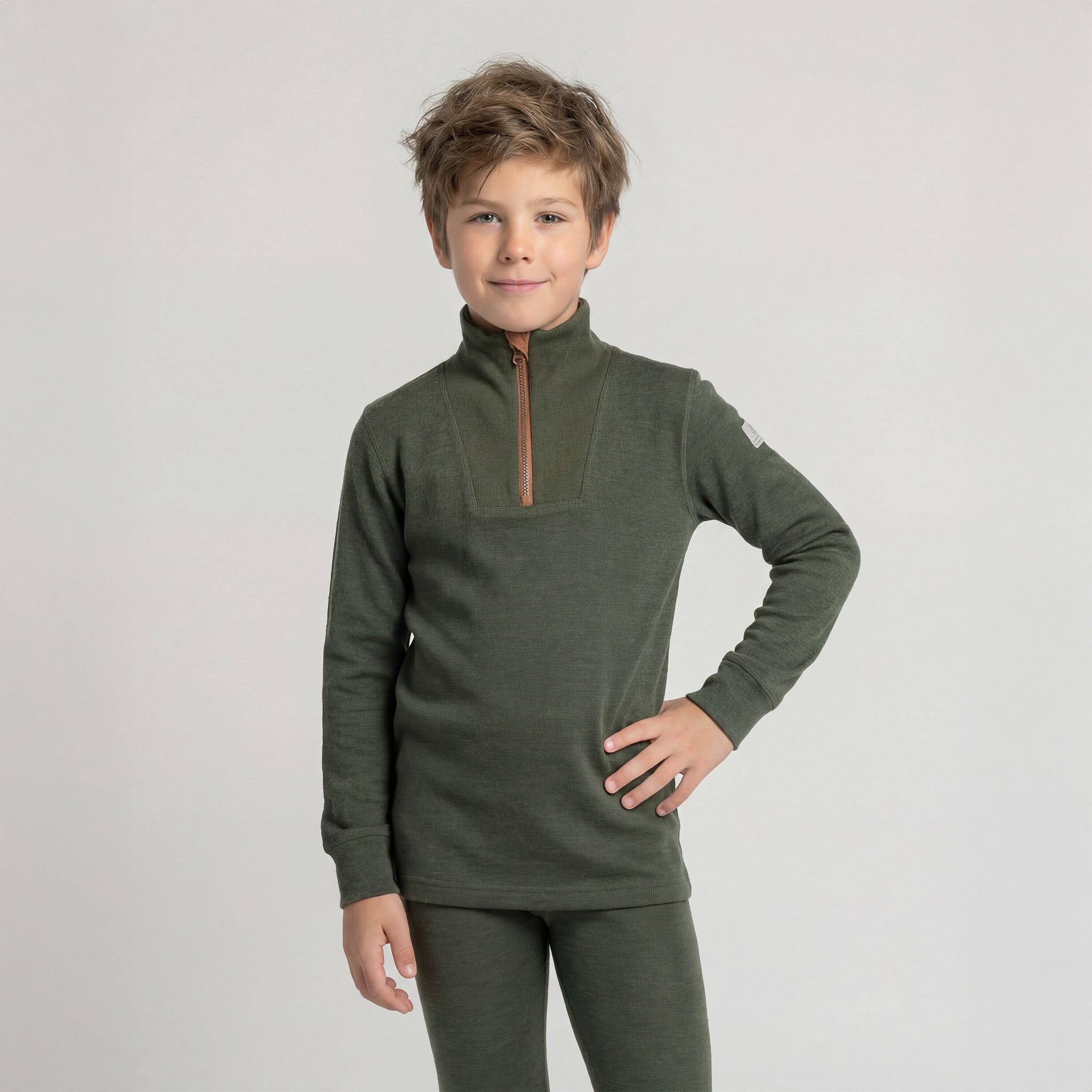 A young boy with light brown hair smiles against a light gray background, wearing the Janus Wool Janus Go 250 Kids Polo with zip merino and matching pants for a cozy, stylish mid-layer look.