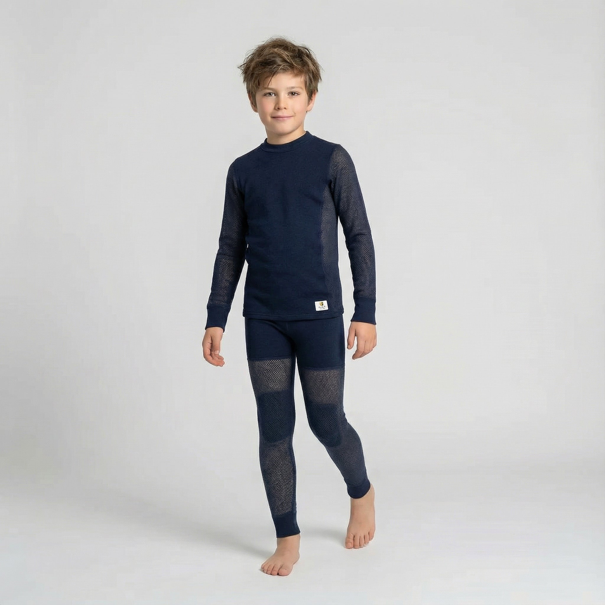 A young boy with tousled hair stands barefoot against a plain light background, dressed in the Janus Wool Jersey mesh merino - Janusull 165 Kids matching navy blue long-sleeve thermal top and leggings.