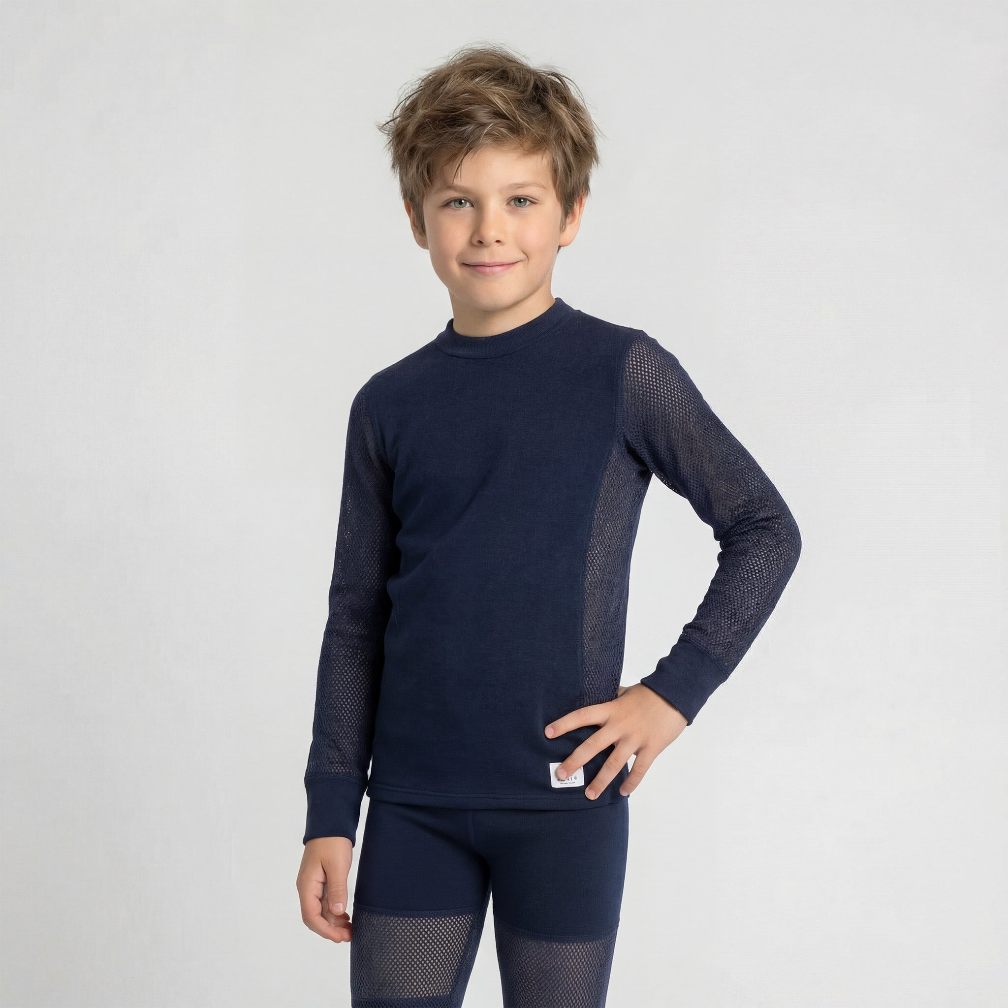A young boy with tousled hair stands confidently against a plain light background, wearing the Janus Wool Jersey mesh merino - Janusull 165 Kids set: a navy blue merino wool long-sleeve top and leggings with mesh panels on sleeves and sides.