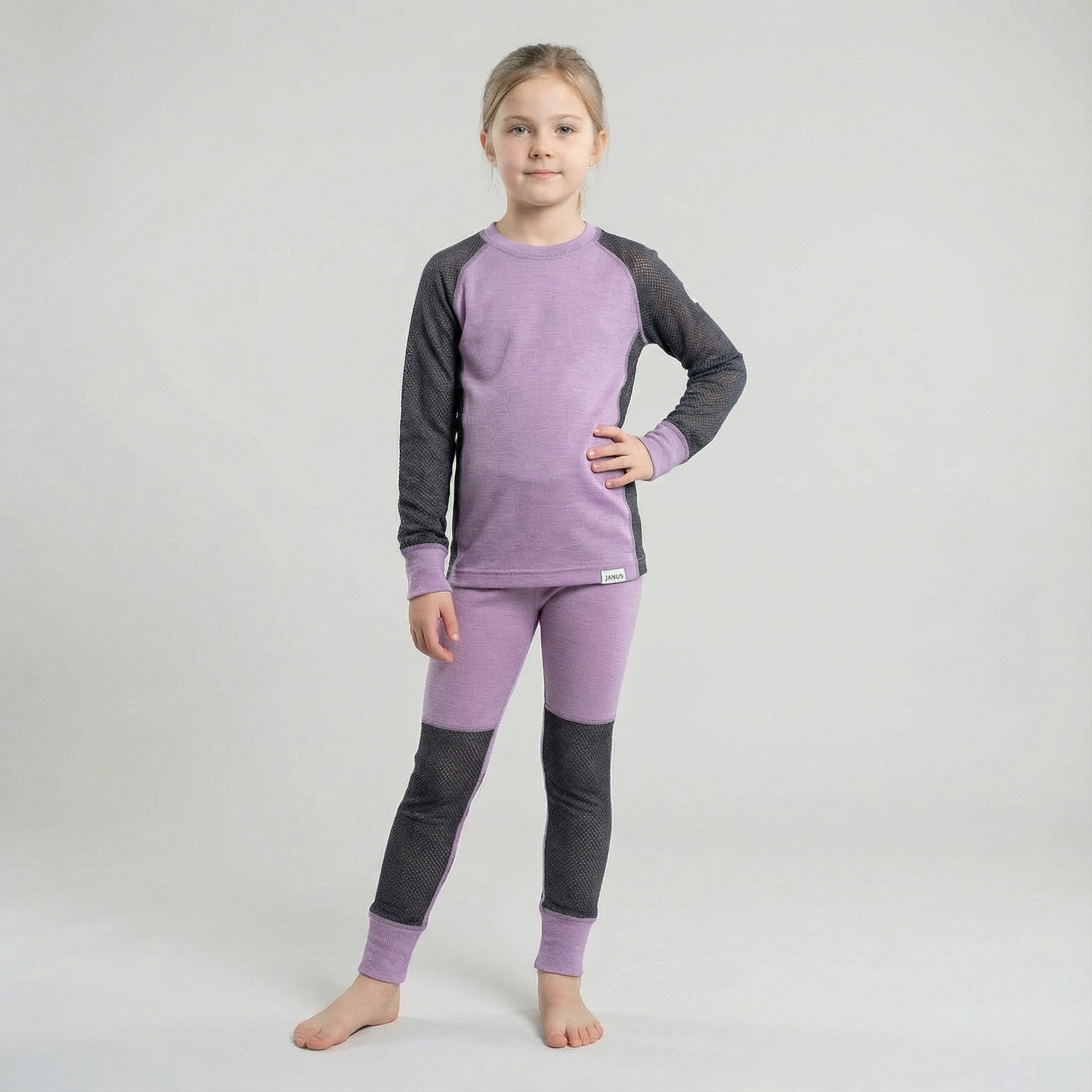 A young girl stands barefoot on a light grey background, wearing purple Leggings mesh merino - Janusull 165 Kids and a matching long-sleeve top with dark grey sleeves and knee patches. She has her left hand on her hip and a neutral expression.
