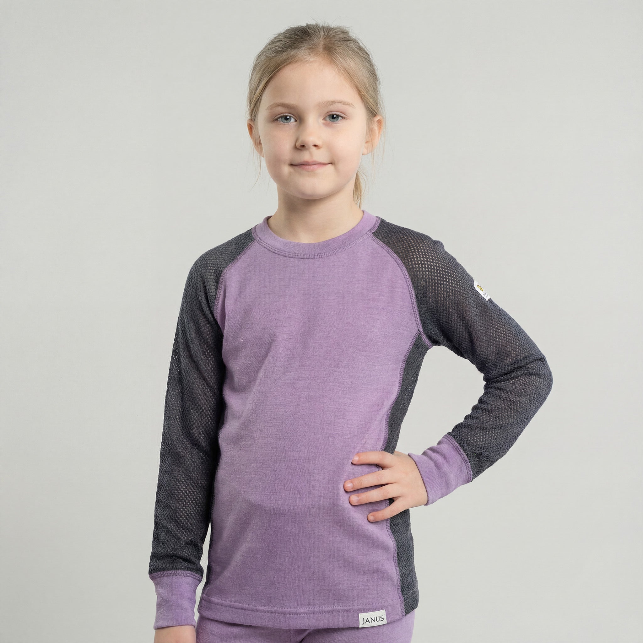 A young girl with light brown hair in a ponytail stands against a plain gray background, wearing the Janus Wool Jersey mesh merino - Janusull 165 Kids long-sleeve top, hand on hip, looking calmly at the camera.