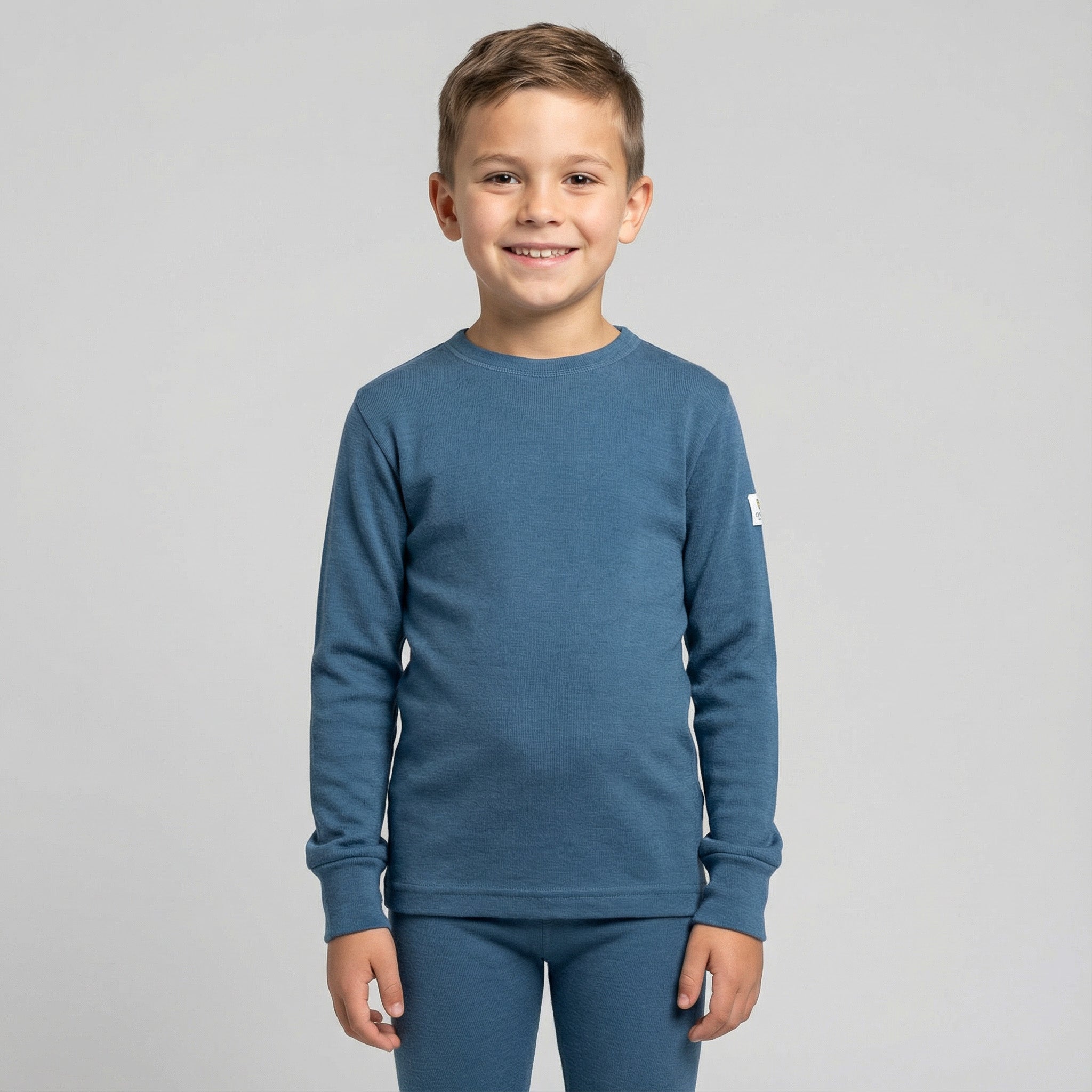 Smiling young boy with short brown hair wears the Jersey merino - Janusull 180 Kids blue baselayer, standing against a plain light gray background.
