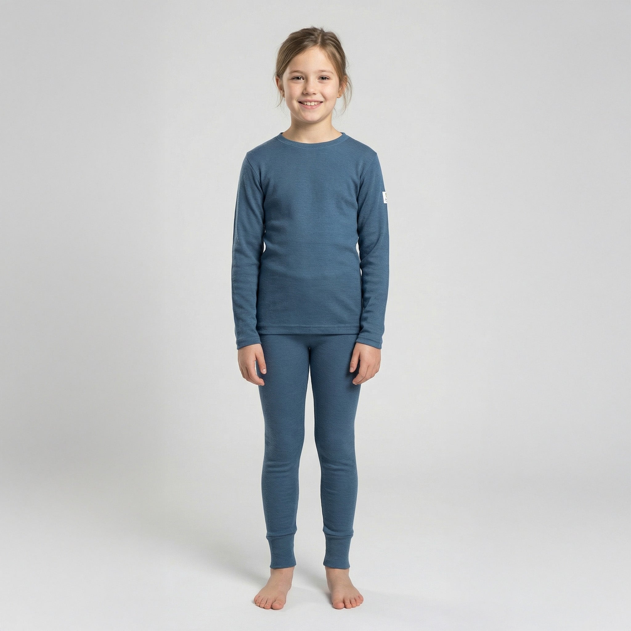 A young girl stands barefoot on a light background, smiling and wearing the Jersey merino - Janusull 180 Kids: a matching blue long-sleeve thermal top and leggings set.